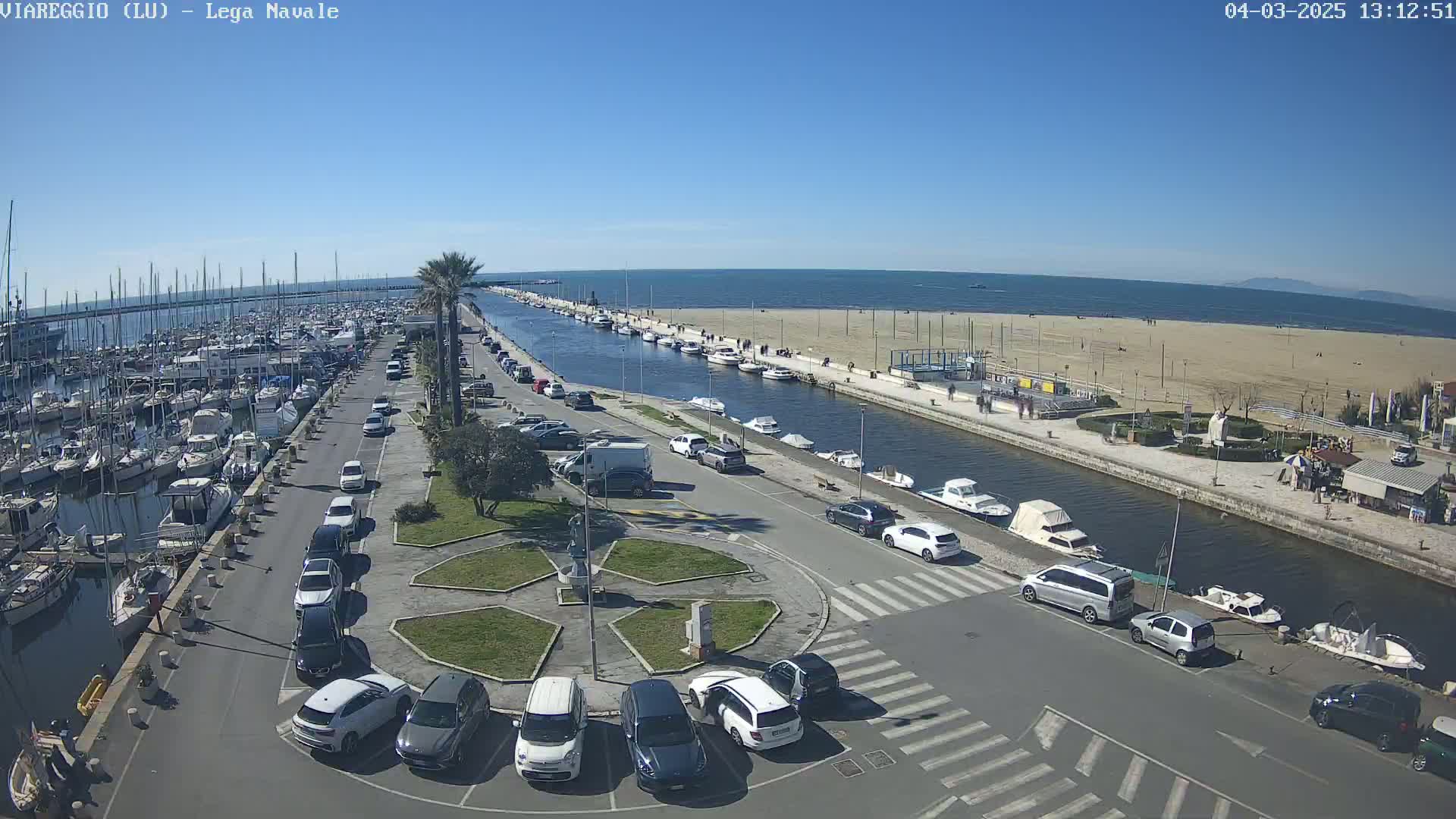 A sunny day shows a marina filled with boats, a canal lined with parked cars and small boats, and a sandy beach beyond.