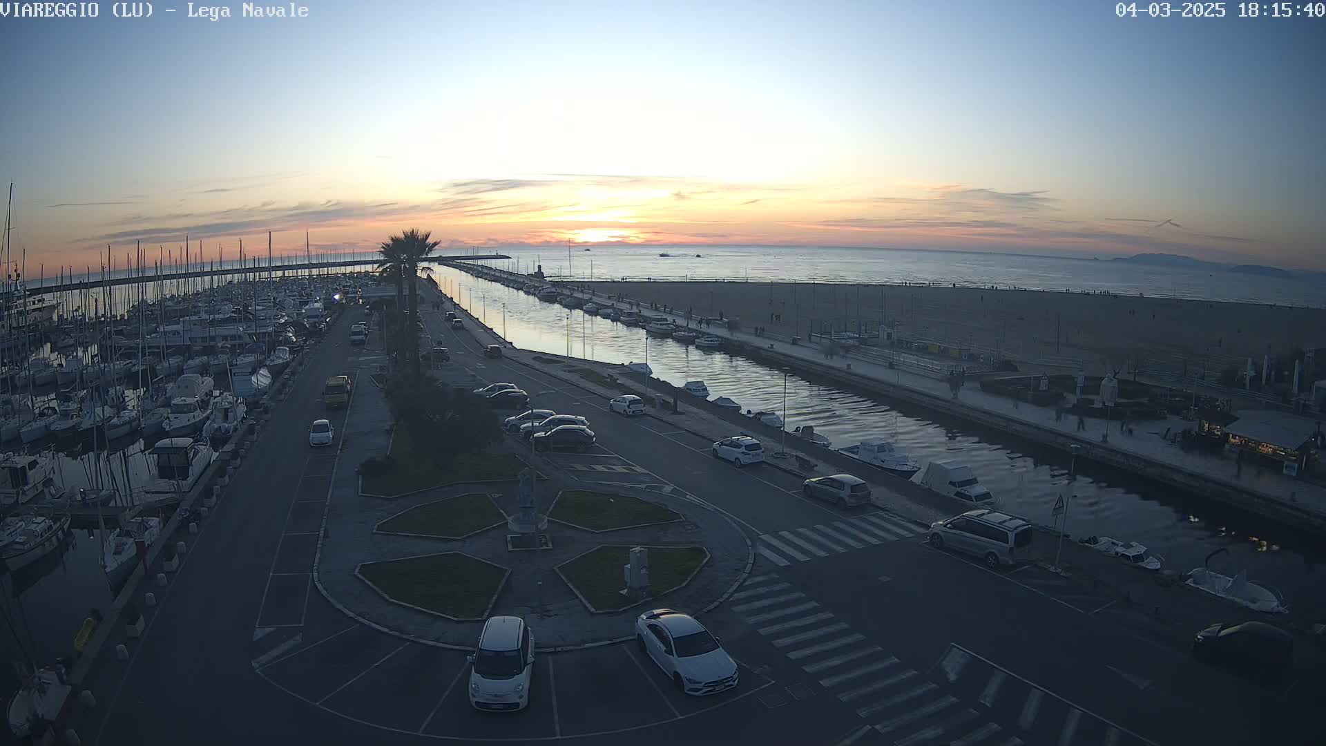 A harbor filled with boats at sunset, next to a beach with people walking along the shore, with cars parked along a canal-side road.