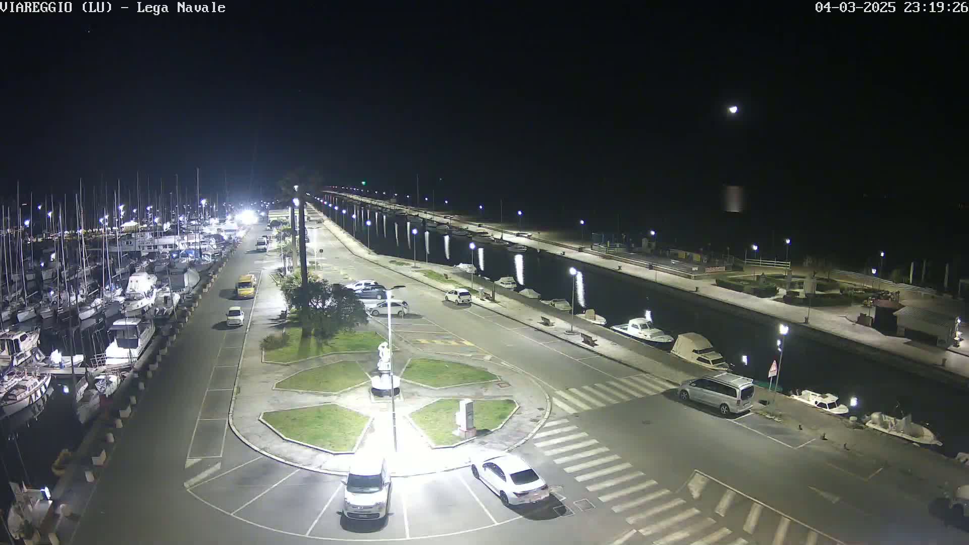 A nighttime, high-angle view shows a marina filled with boats, a paved area with cars parked, and a small roundabout with a statue in the center.
