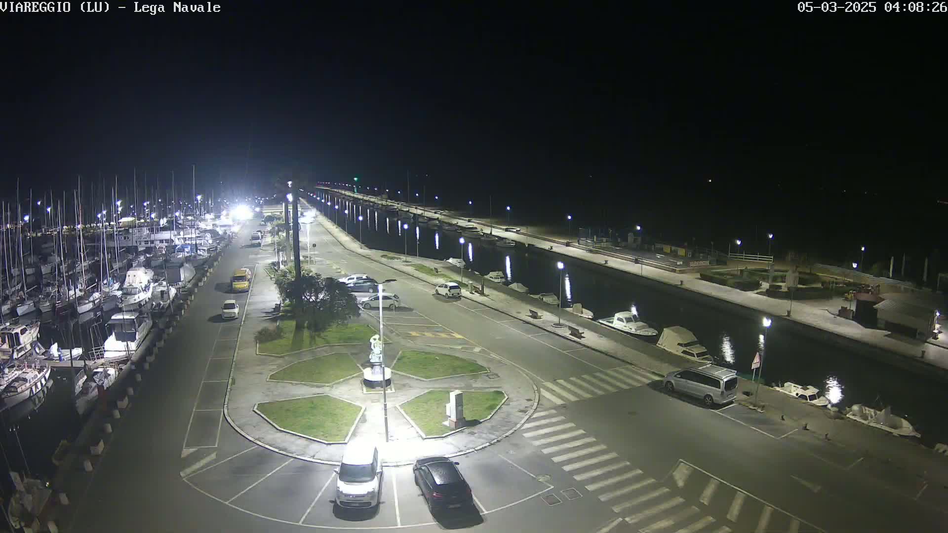 A nighttime aerial view of a marina with numerous boats docked, a roadway with parked cars, and a circular plaza in the foreground.