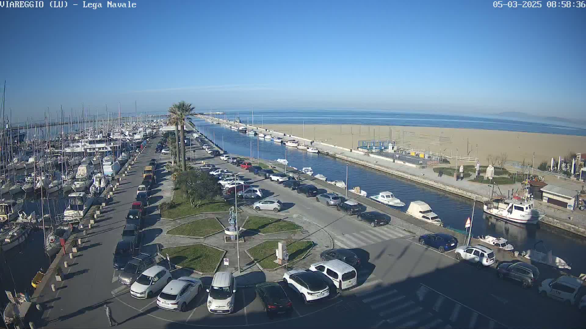 A marina filled with boats is adjacent to a sandy beach and calm ocean under a clear blue sky.