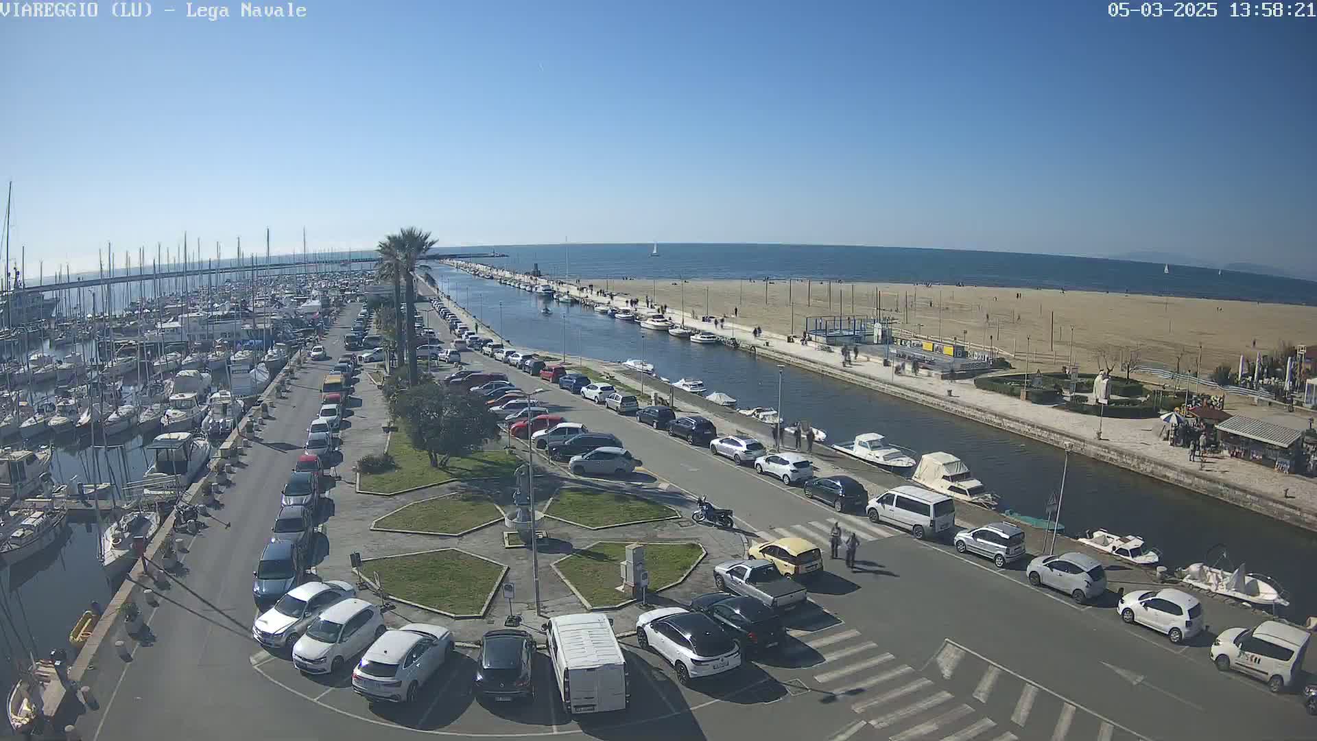 A marina filled with boats is next to a sandy beach and calm ocean under a clear blue sky, with cars parked along a canal separating the marina from the beach.