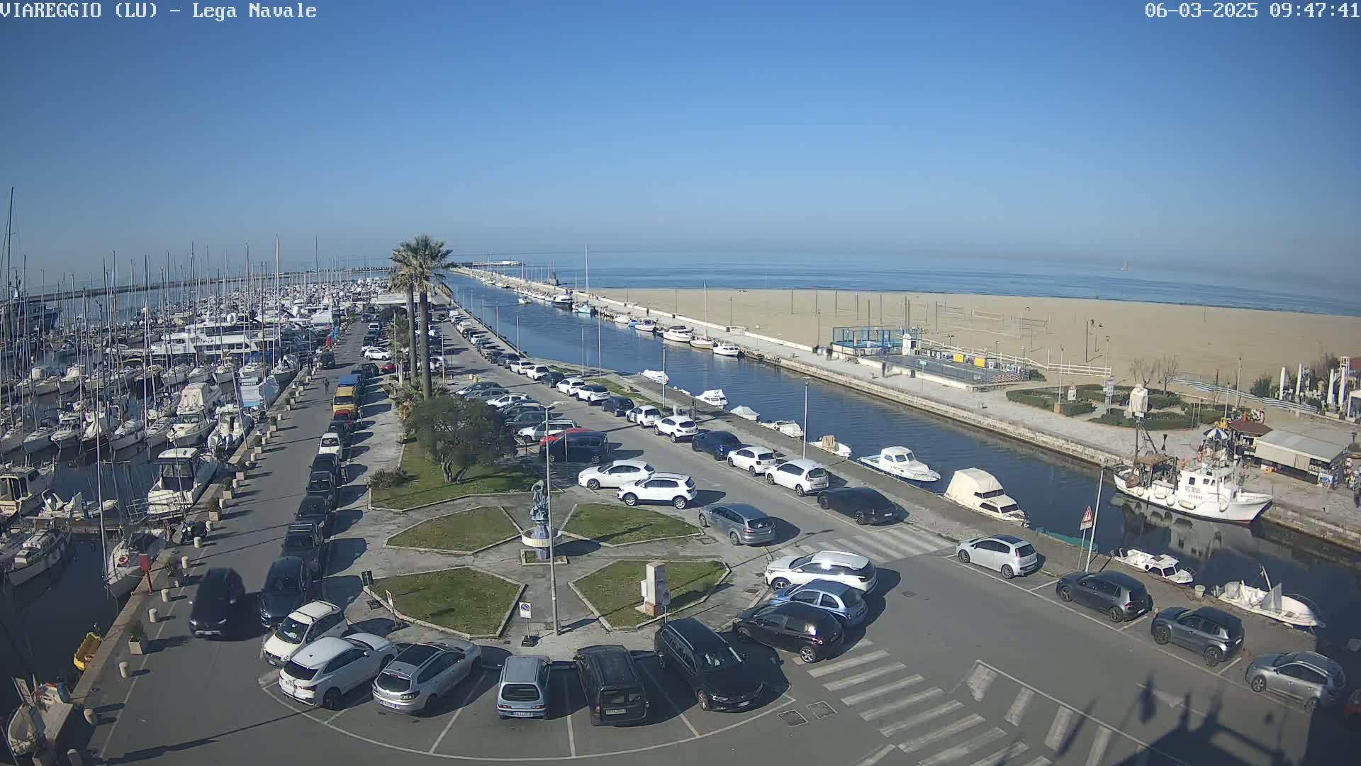 A harbor filled with boats is adjacent to a sandy beach and a parking lot filled with cars under a clear, sunny sky.