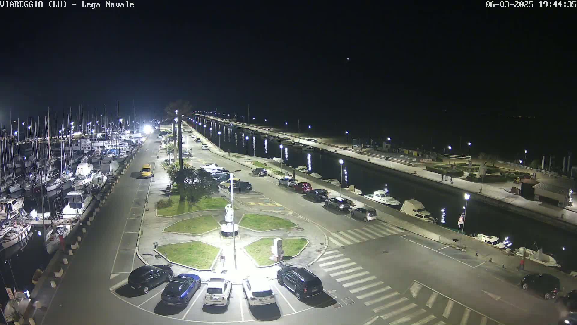 A nighttime, aerial view of a marina with numerous boats docked, a parking lot with cars, and a paved walkway along a calm waterway.