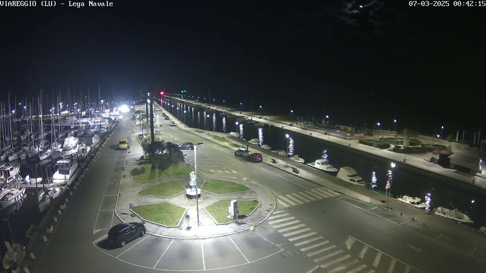 A nighttime, illuminated view of a marina with numerous boats docked alongside a canal, and a roadway with parked cars leading to a small, landscaped roundabout.