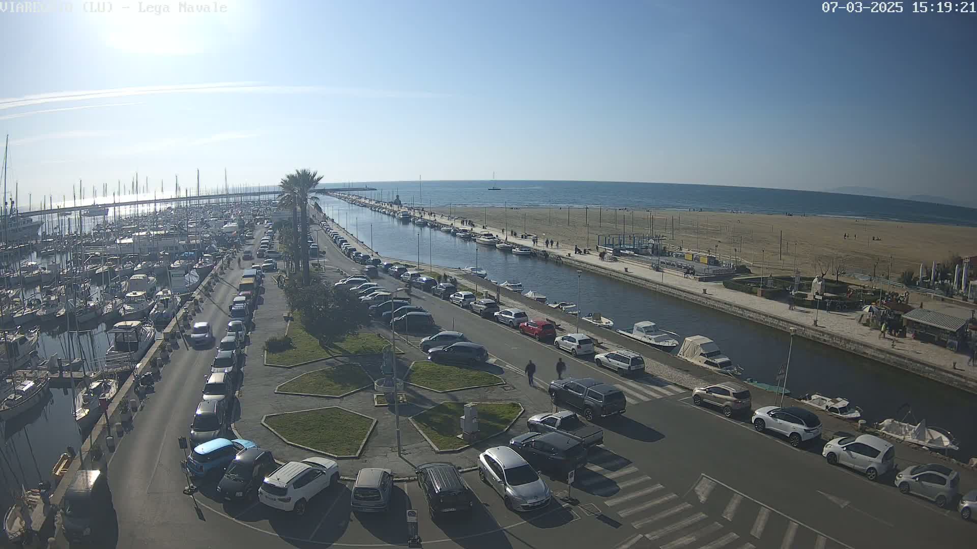 A sunny day at a harbor shows a marina filled with boats, a canal lined with parked cars and people, and a sandy beach beyond.