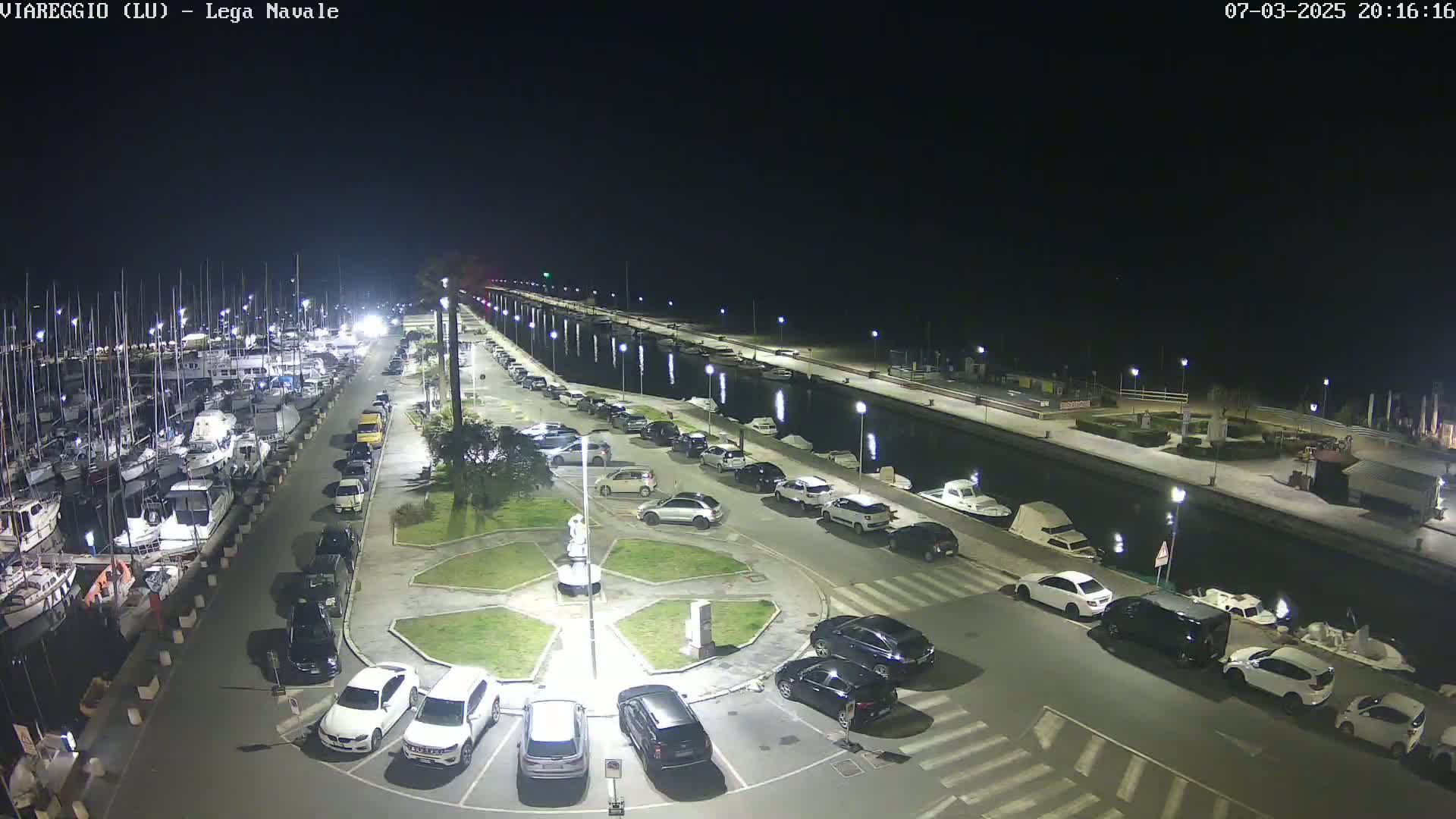 A nighttime aerial view of a marina with numerous boats docked, a parking lot filled with cars surrounding a small plaza, and calm water.