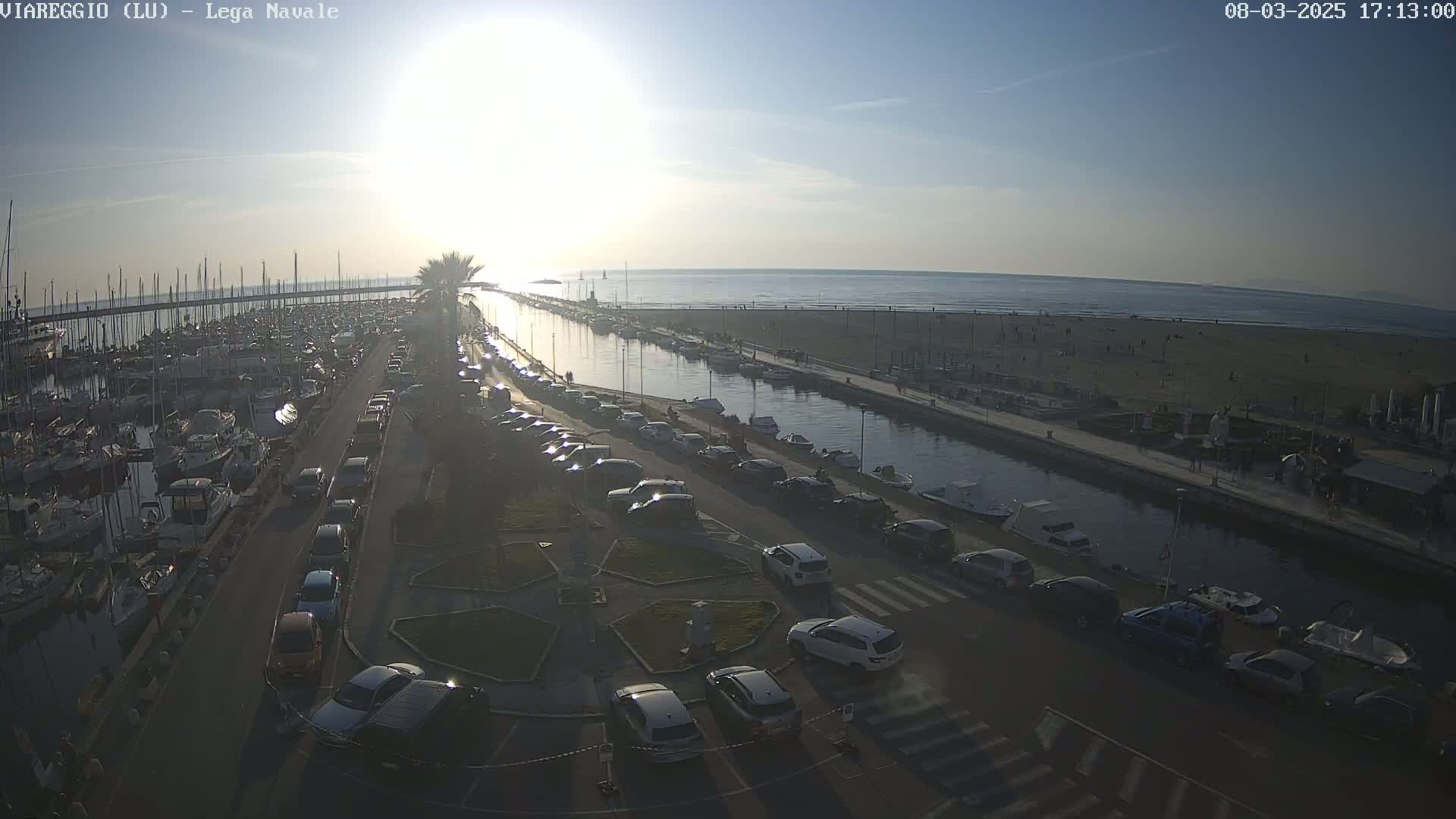 A marina filled with boats is next to a canal that leads to a beach under a sunny sky.