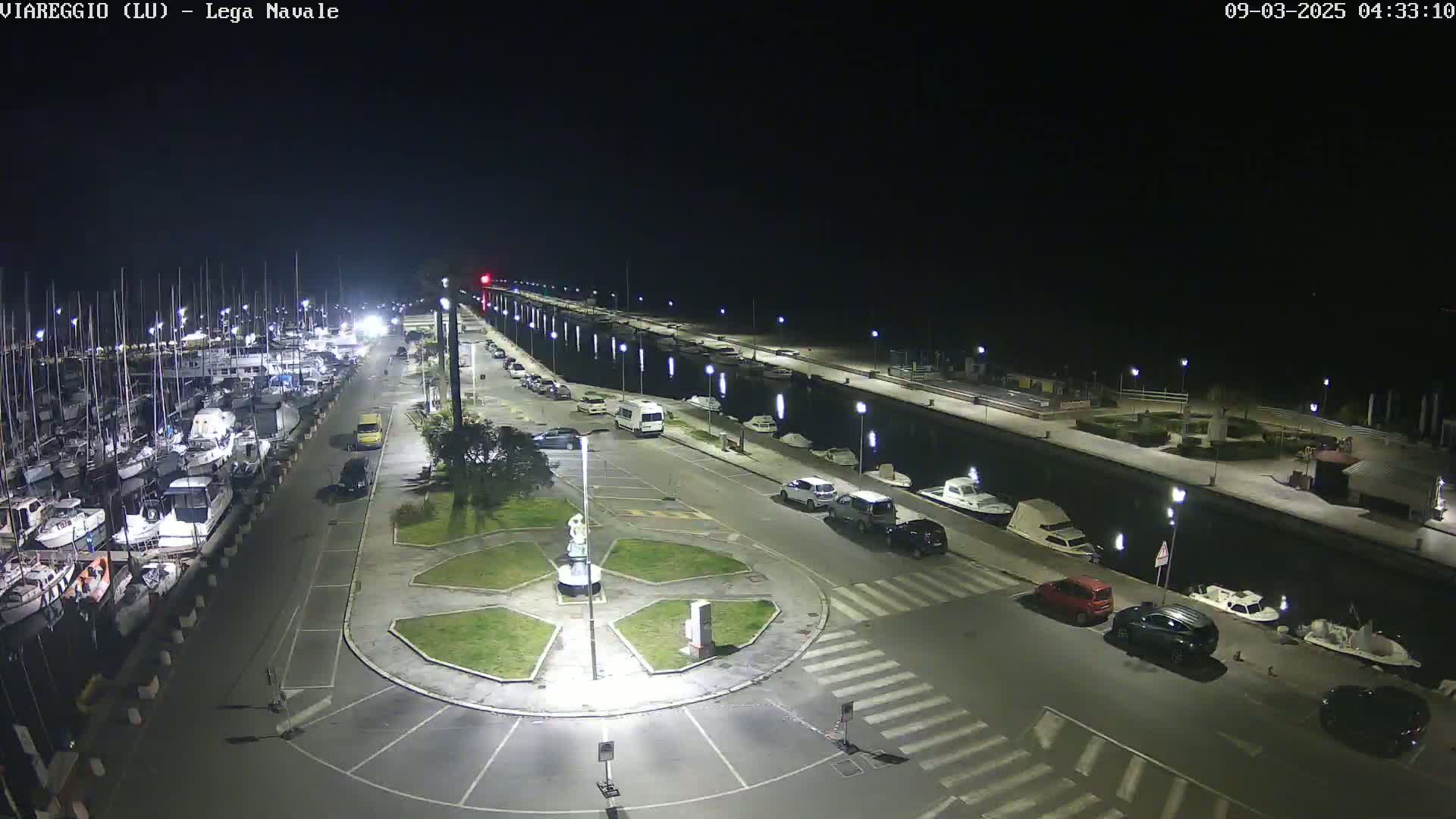 A nighttime aerial view of a marina with numerous boats docked alongside a paved waterfront area containing parked cars and a small central plaza.