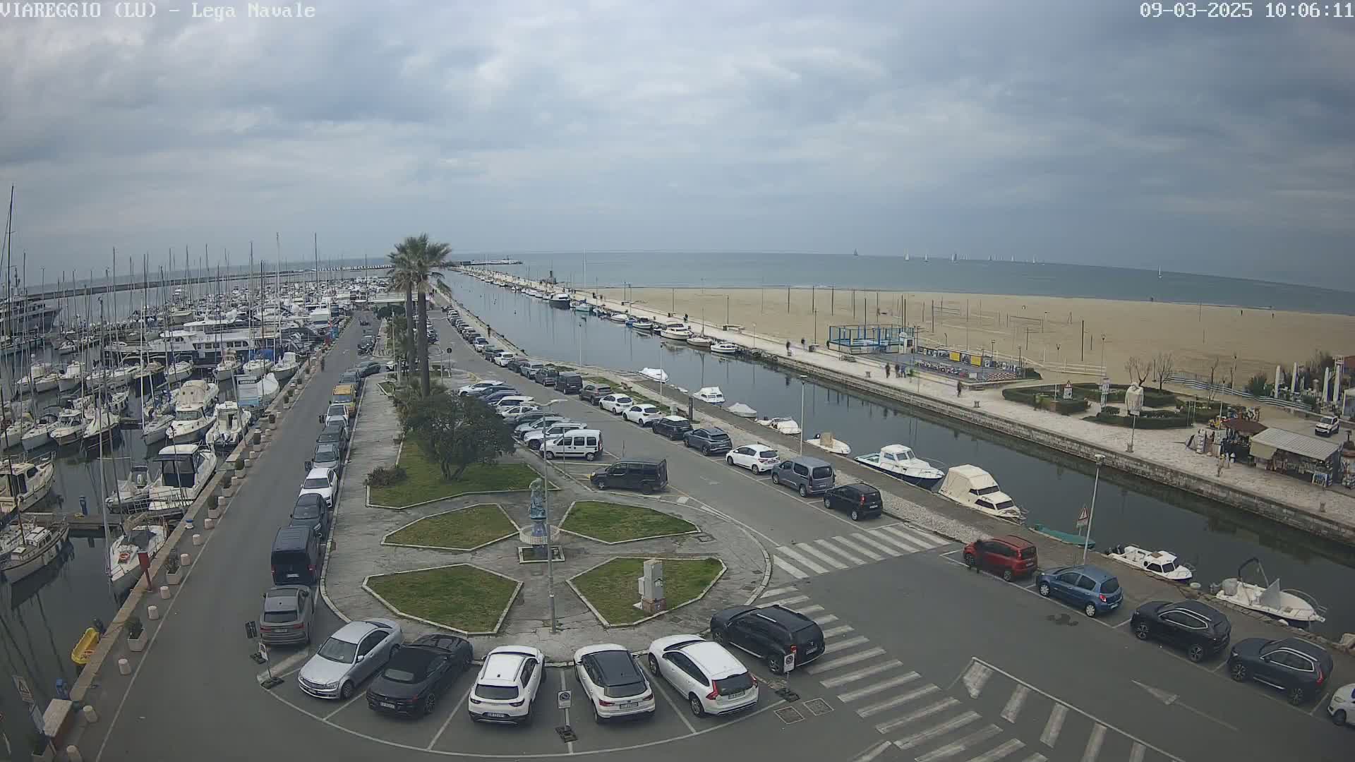 A marina filled with boats is next to a canal and sandy beach under a cloudy sky.