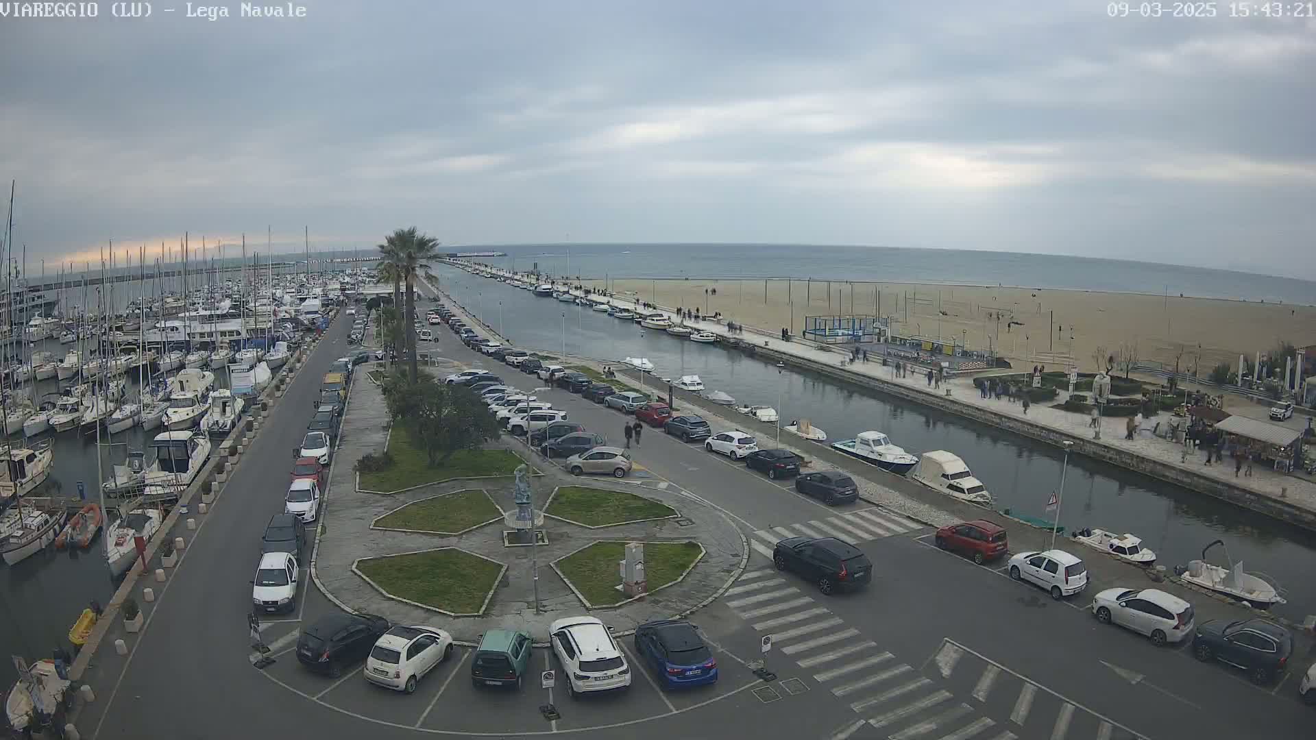 An overcast day shows a marina filled with boats, a canal with additional boats and pedestrians on the shoreline, and a parking lot full of cars surrounding a small circular plaza.