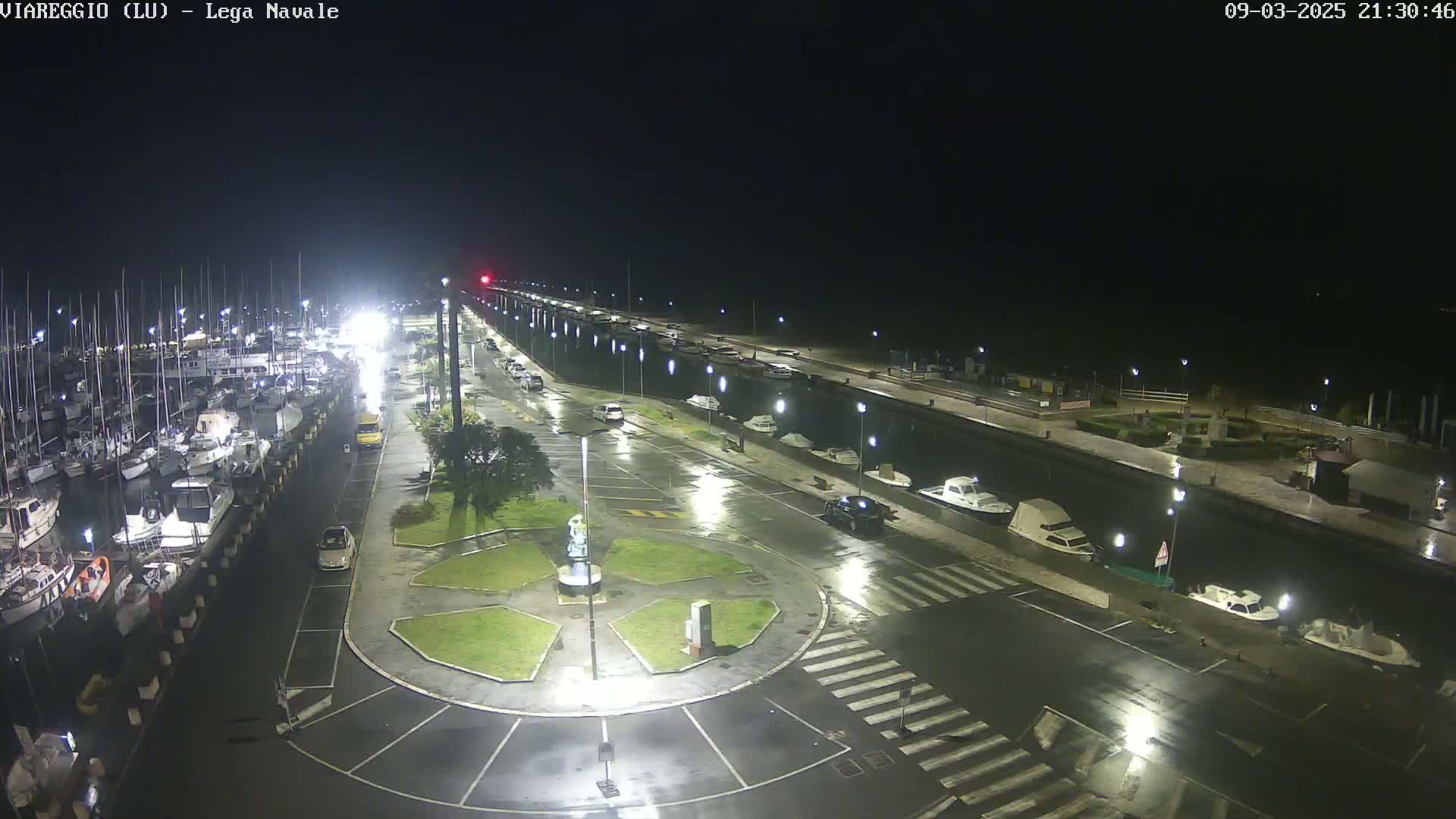 A nighttime, rain-slicked marina with numerous boats docked, surrounded by wet pavement and streetlights, features a small, circular plaza in the center.