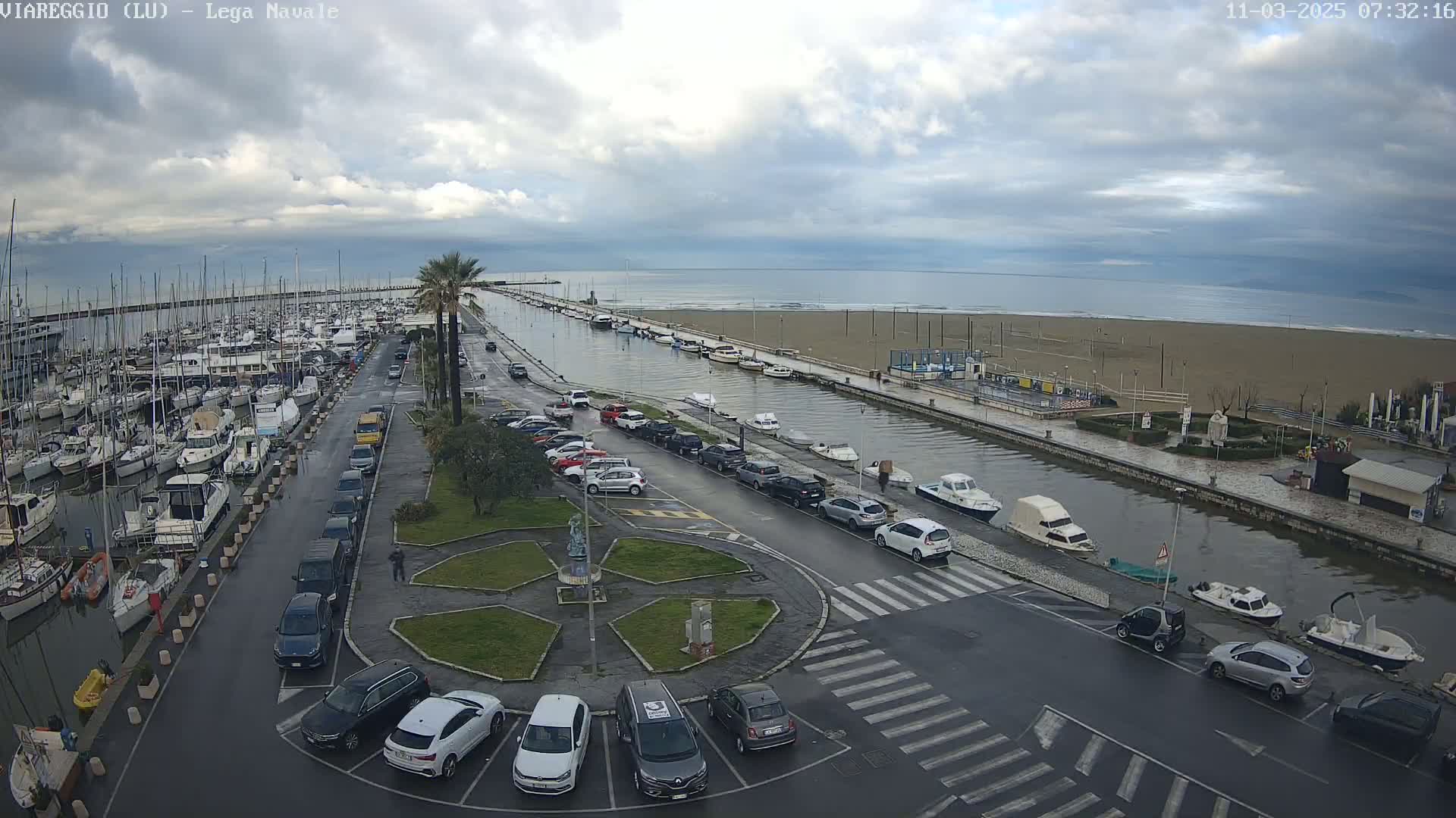 A marina filled with boats is next to a canal, which is partially flooded and alongside a beach under an overcast sky.