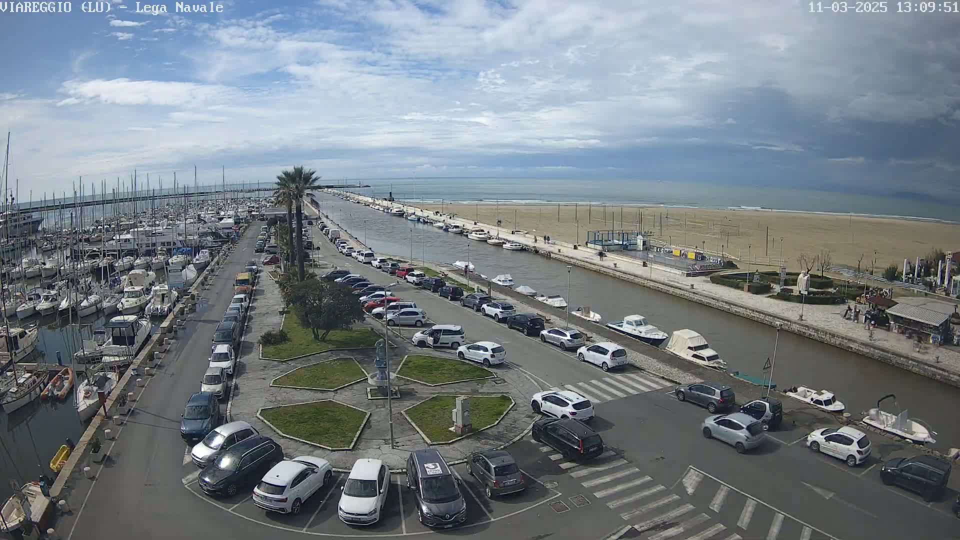 A marina filled with boats is next to a canal and parking lot, all overlooking a sandy beach and ocean under a partly cloudy sky.