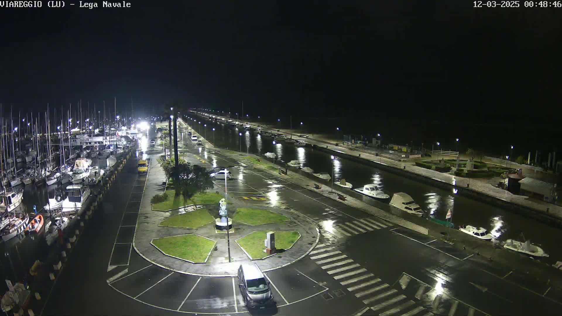 A wet, nighttime view of a marina with numerous boats docked, a canal, and a roadway with a small roundabout, all under dark skies.