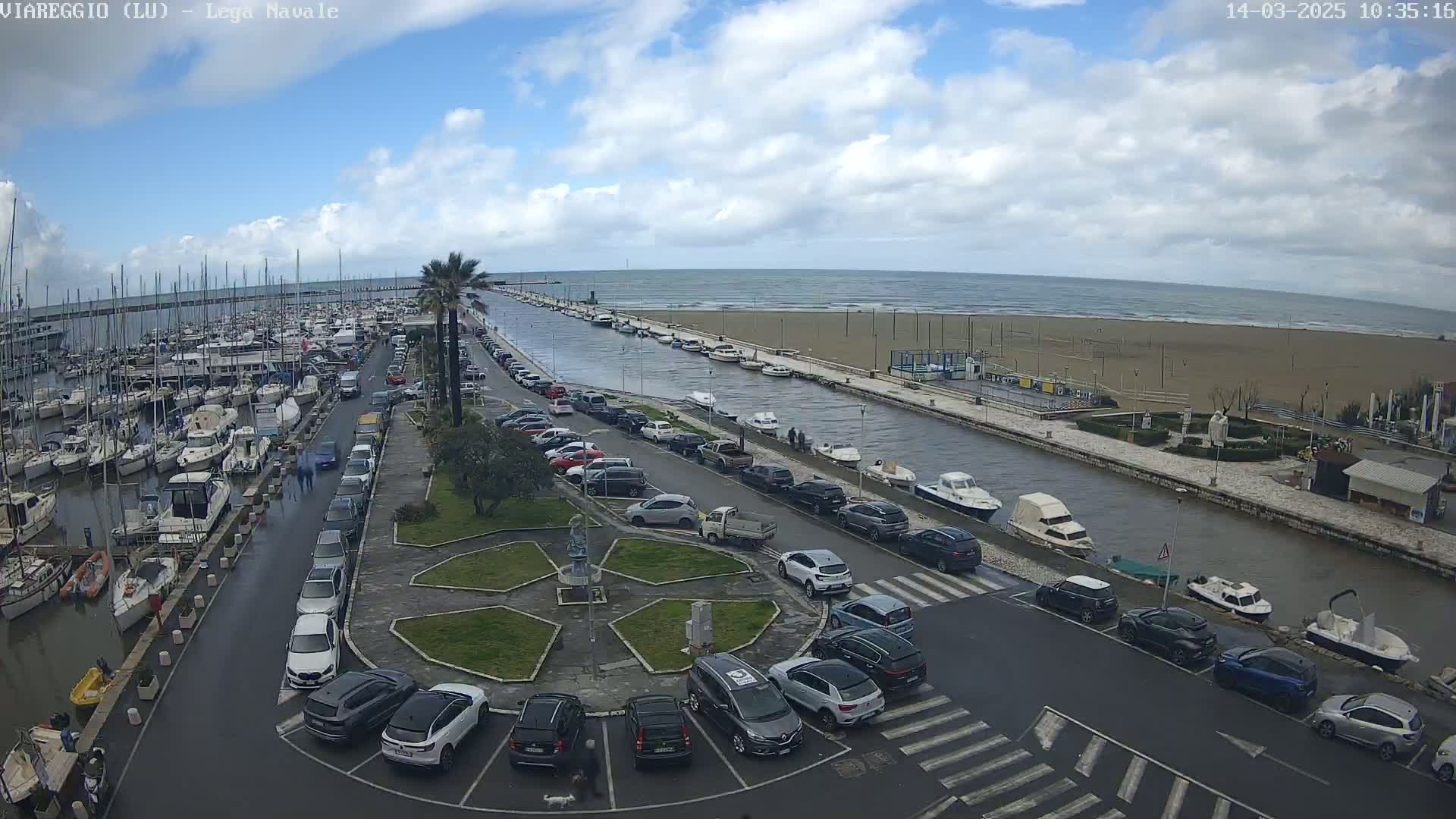A harbor filled with boats is next to a canal lined with parked cars, leading to a sandy beach under a partly cloudy sky.