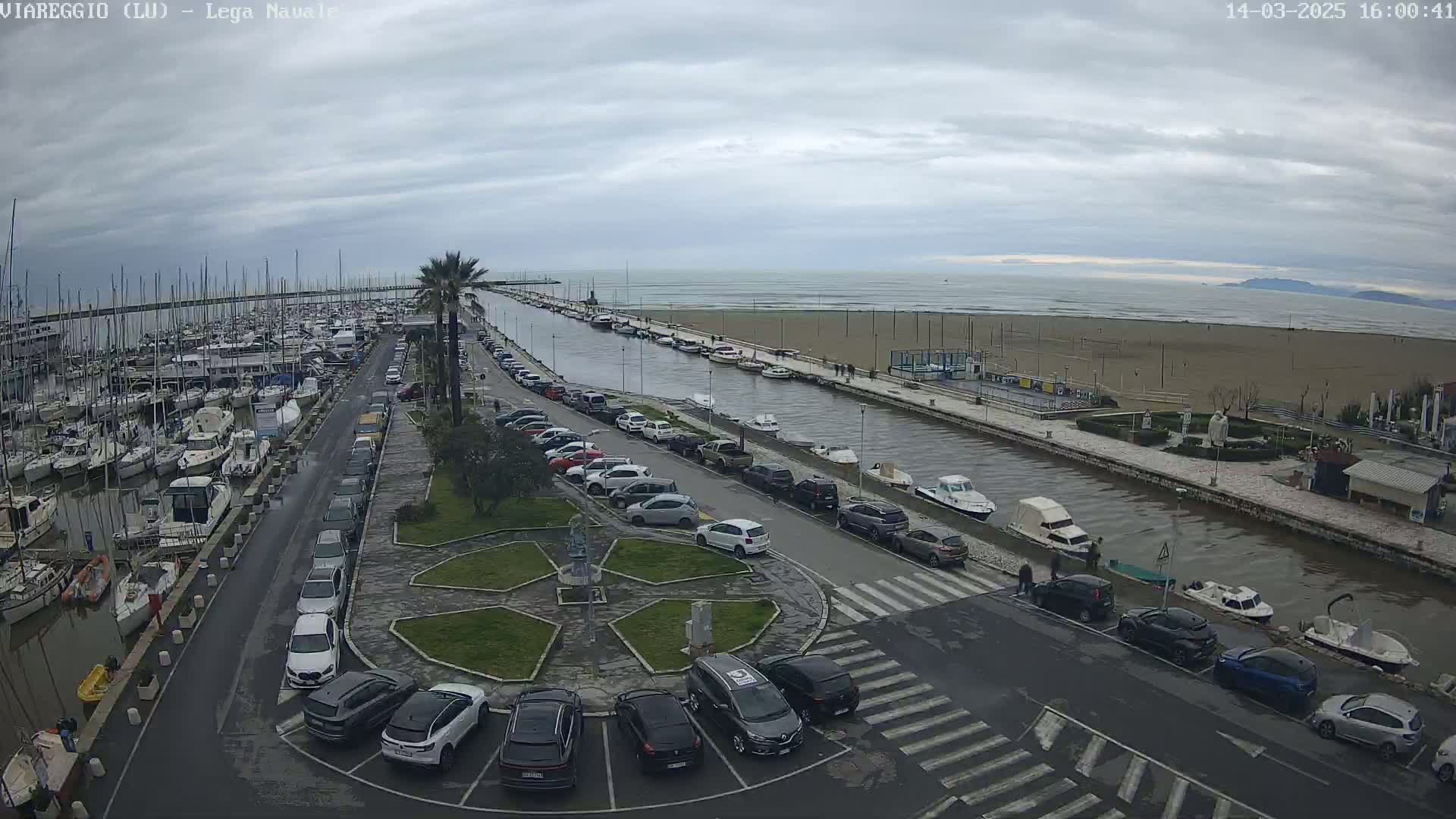 A marina filled with boats is next to a canal lined with parked cars, leading to a sandy beach under a cloudy sky.