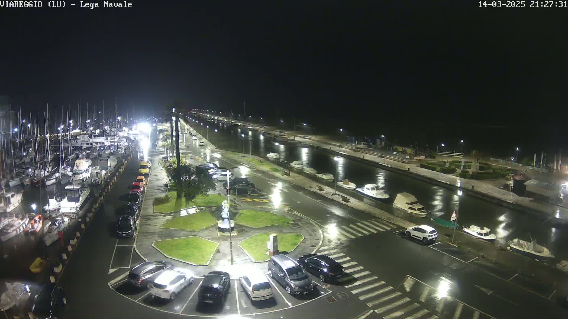A nighttime, rain-soaked marina with docked boats and cars parked along a wet roadway.