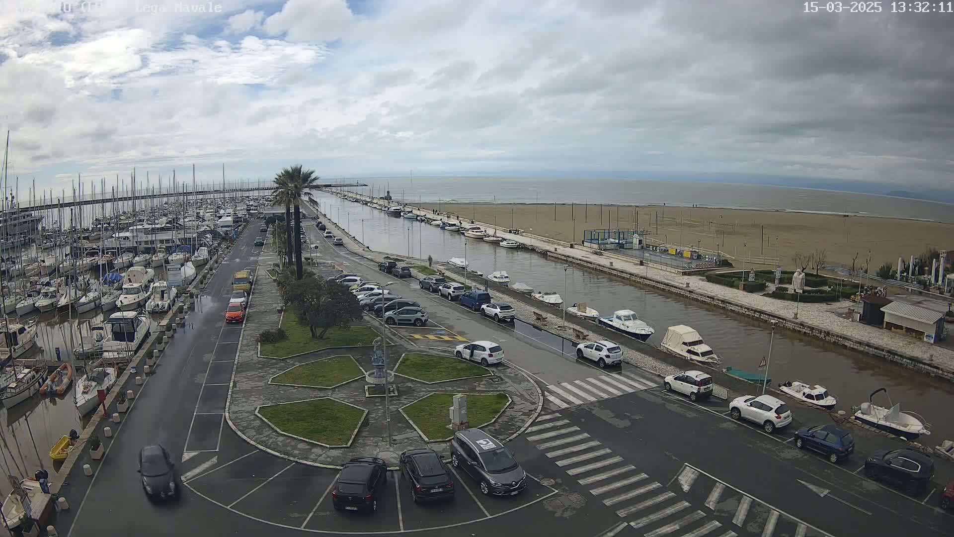 A harbor filled with boats is next to a sandy beach under an overcast sky, with cars parked along a road leading to a central roundabout.