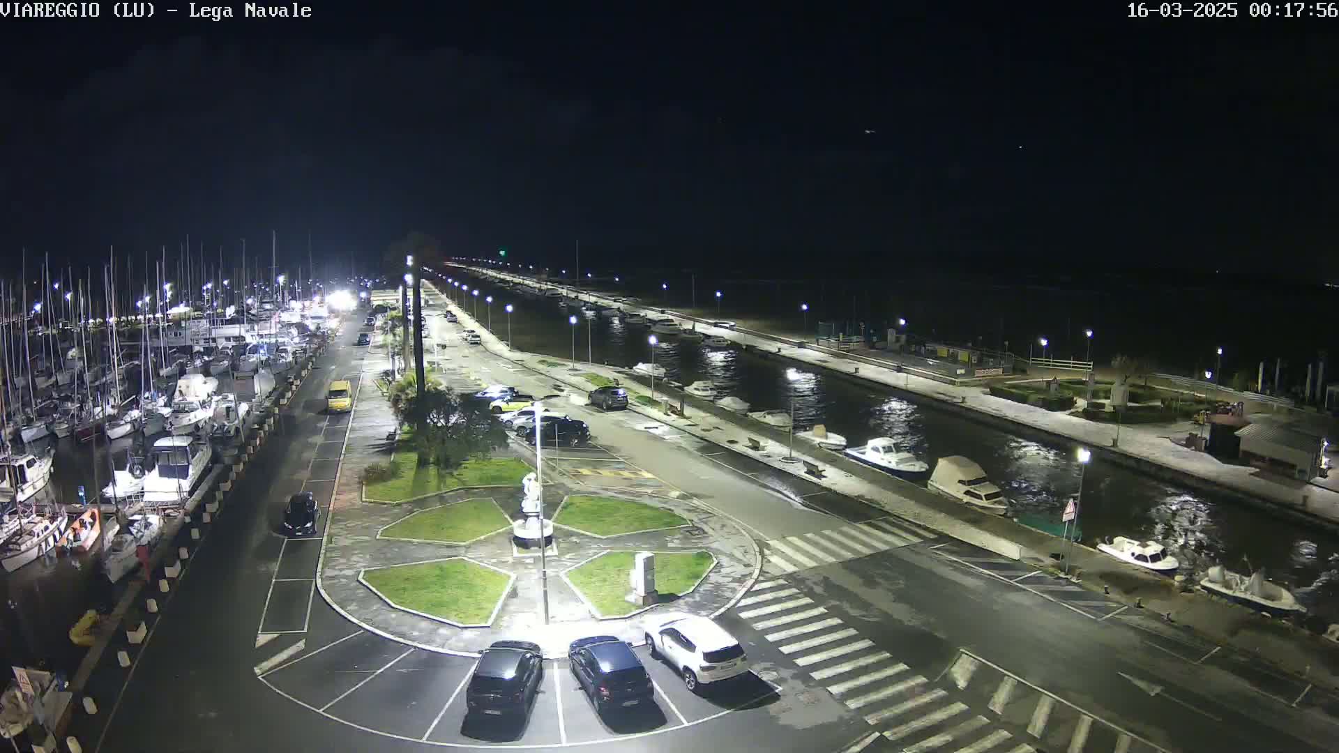 A nighttime, aerial view of a marina with docked boats, a paved area with cars parked, and a small roundabout in the center.