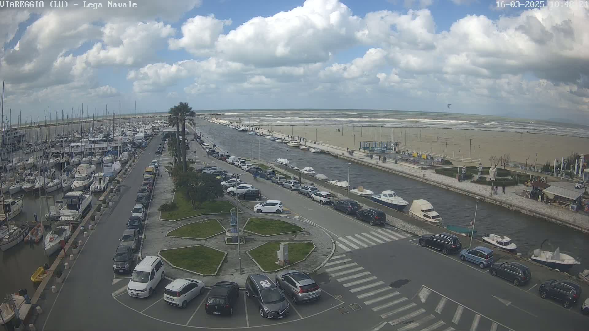 A harbor filled with boats and surrounded by cars parked along a canal leading to a windy, partly cloudy ocean.