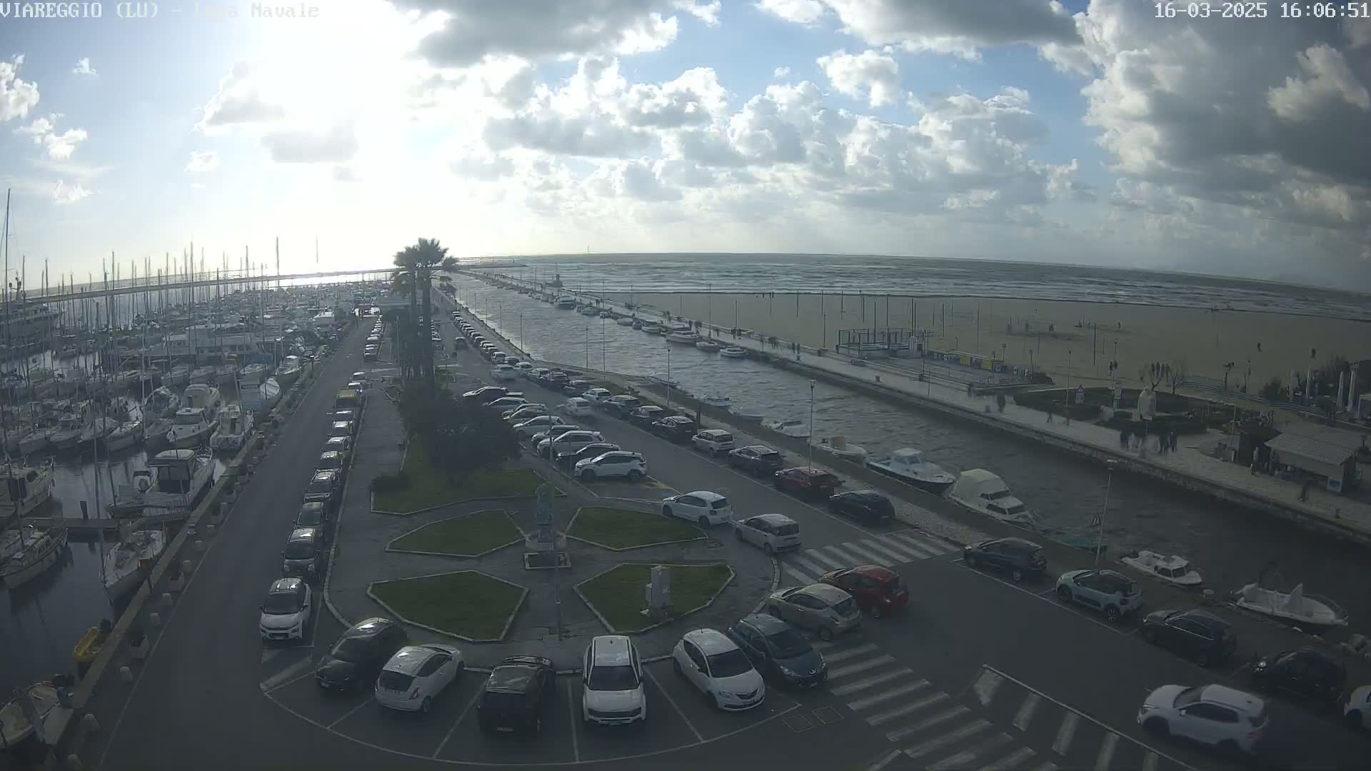 A marina filled with boats is next to a canal and a beach with a partly cloudy sky and a choppy sea.