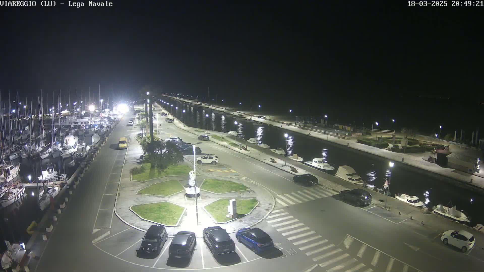 A nighttime aerial view of a marina with numerous boats docked, a paved area with cars parked, and a small landscaped circle in the center.