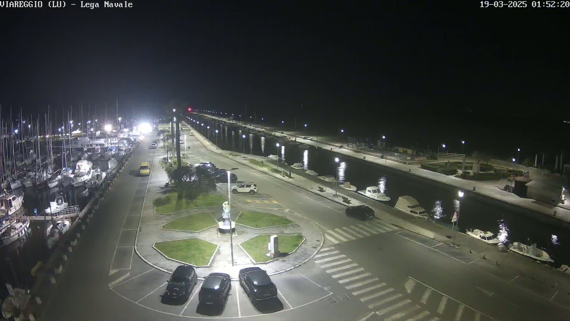 A nighttime view of a marina with docked boats and cars parked in a lot surrounding a central plaza, under clear skies.