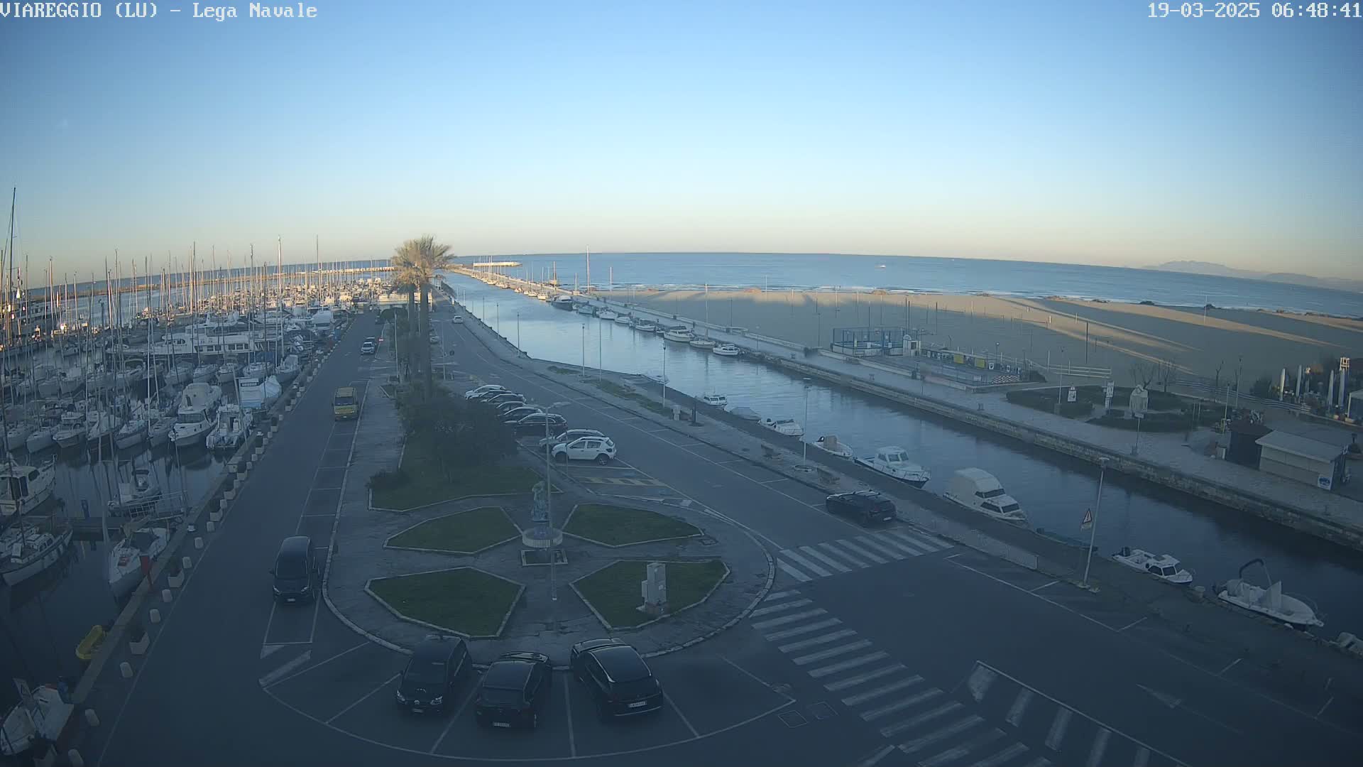 A marina filled with boats is next to a calm, light blue ocean on a clear, sunny day, with a roadway and parking lot separating the water from a sandy beach.