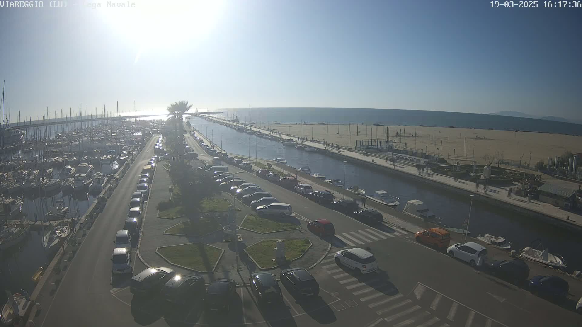 A sunny day overlooking a marina filled with boats, a canal, a parking lot with cars, and a sandy beach beyond.