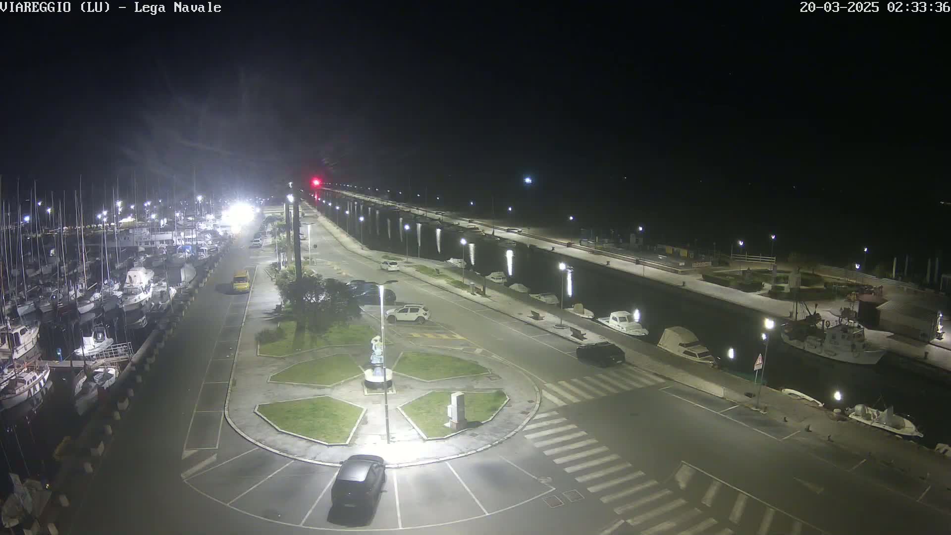 A nighttime view of a harbor filled with boats, a roadway with cars, and a small roundabout in the center, all under a clear, dark sky.