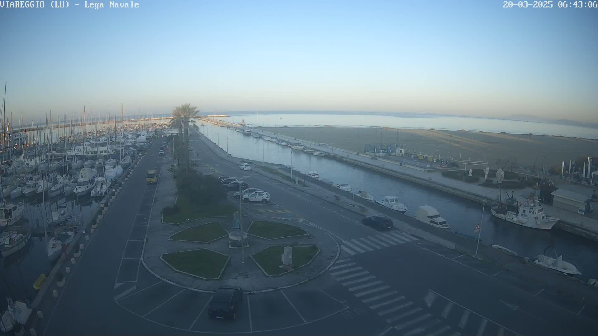 A marina filled with boats is situated next to a calm, wide canal that leads to a sandy beach under a clear, bright morning sky.
