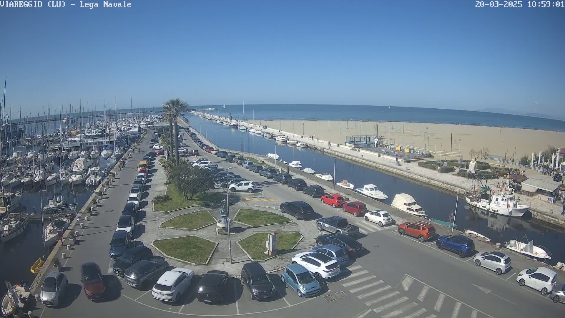 A sunny day overlooking a harbor filled with boats, a canal, a sandy beach, and a parking lot full of cars.