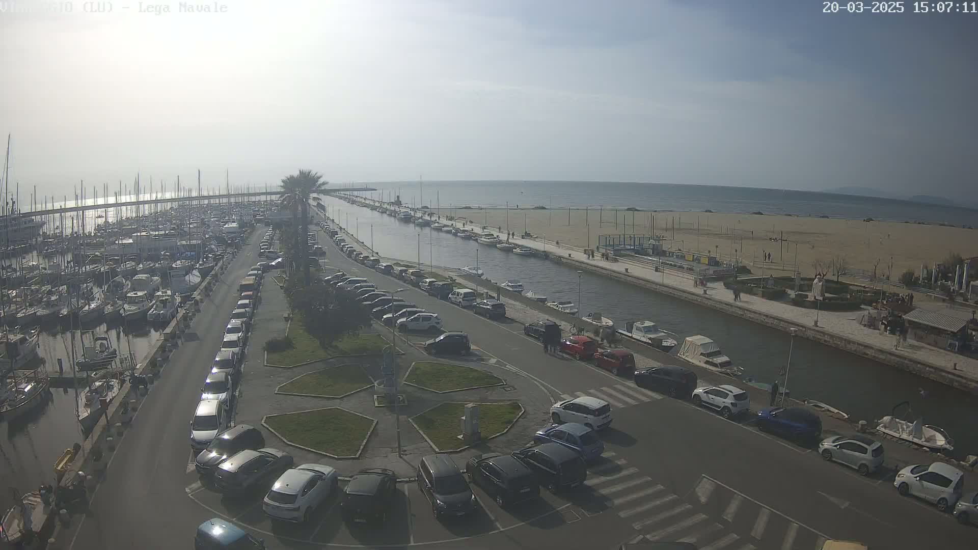 A harbor filled with boats and a sandy beach are visible from an elevated vantage point on a sunny day, with cars parked along a canal separating the two.