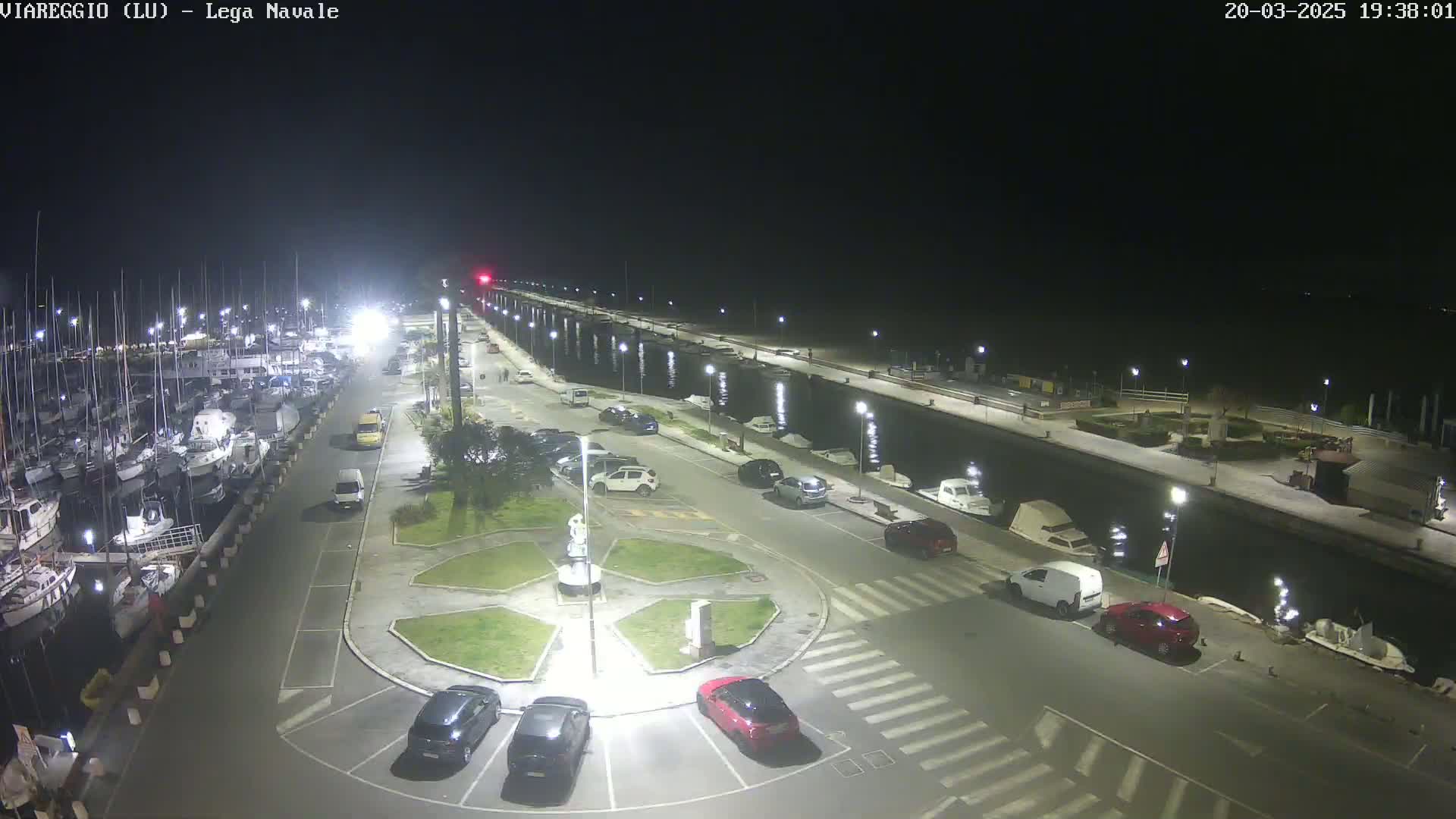 A nighttime aerial view of a marina with docked boats, cars parked in a lot surrounding a small roundabout, and a calm waterway.