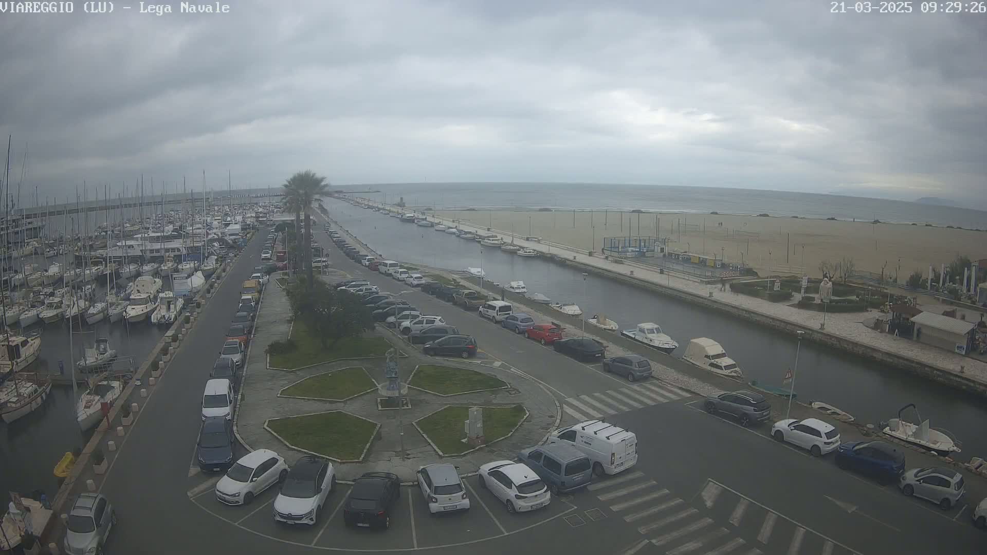 A marina filled with boats is adjacent to a canal that leads to a sandy beach under an overcast sky.