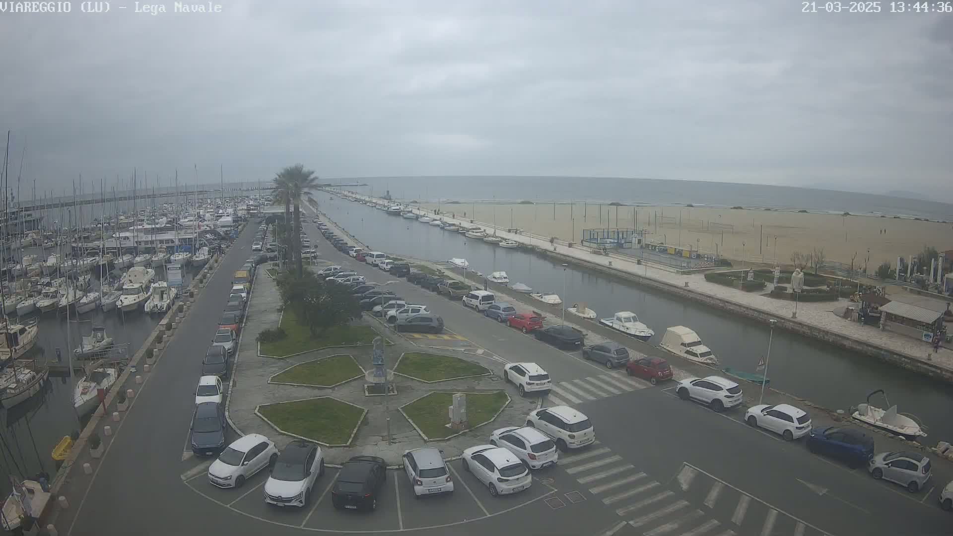 An overcast day reveals a marina filled with boats, a canal lined with parked cars leading to a sandy beach and calm ocean.