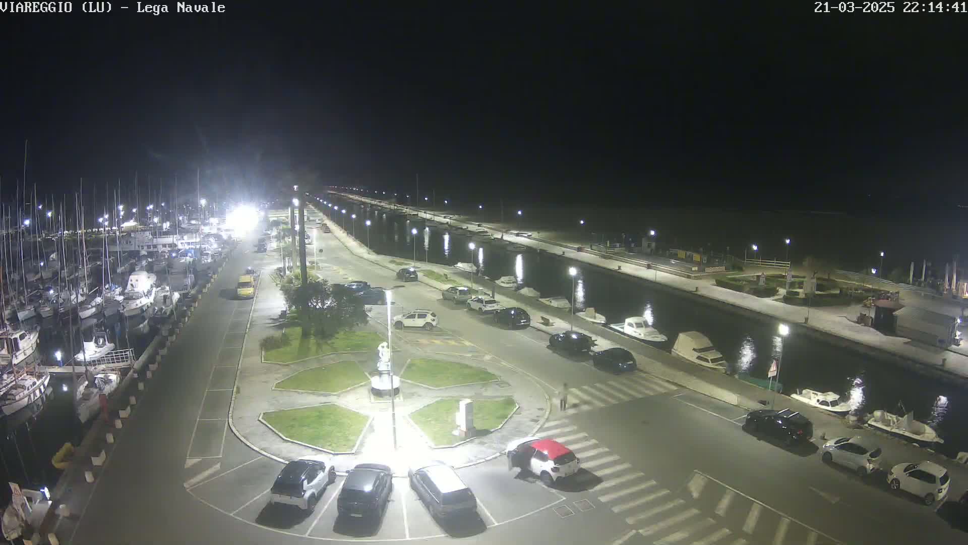 A nighttime view of a marina with numerous boats docked, a roadway lined with parked cars, and a central plaza featuring a statue, all illuminated by artificial lights.