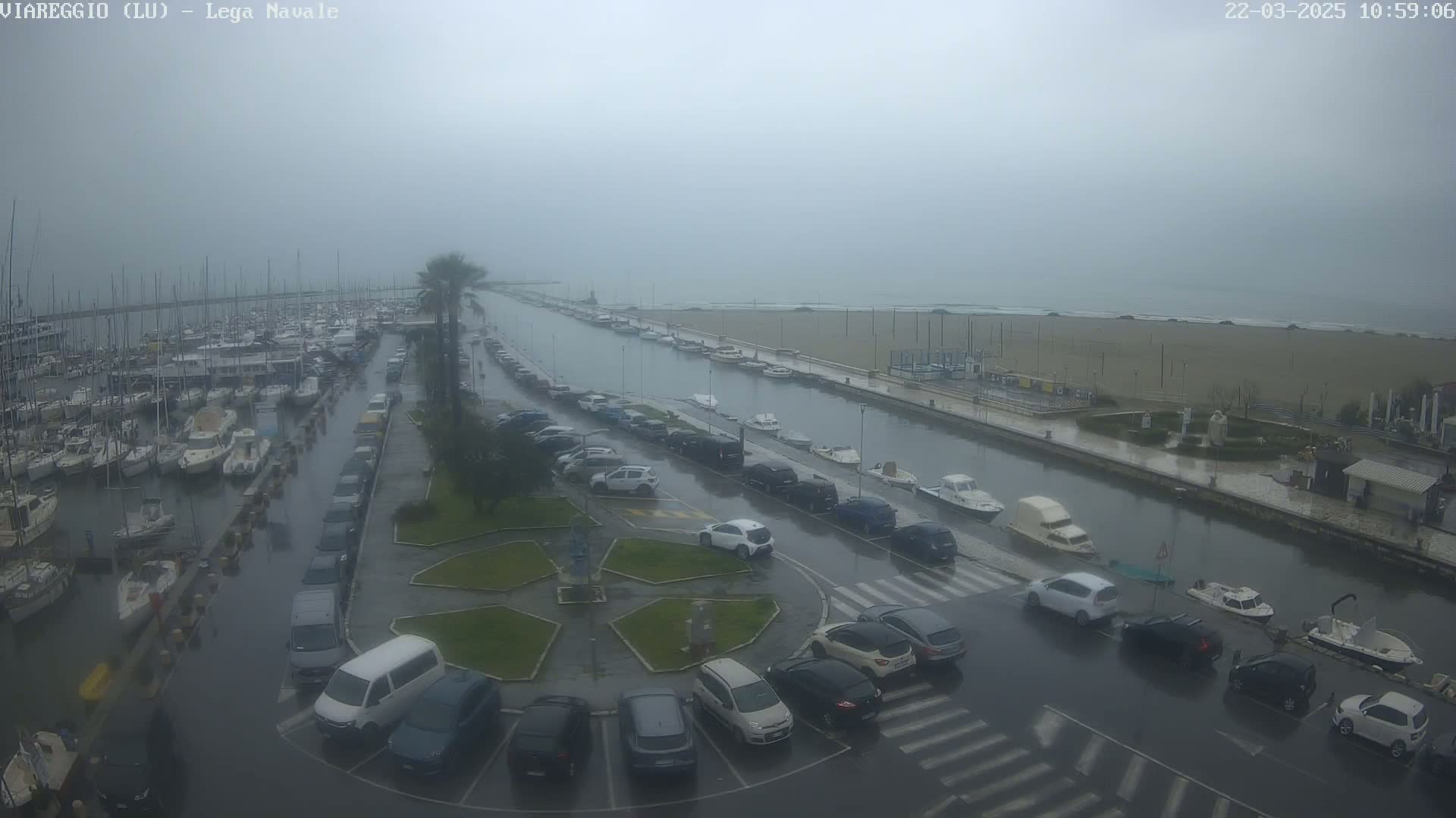 A rain-swept marina with numerous boats and cars parked along a canal leading to a beach under an overcast sky.