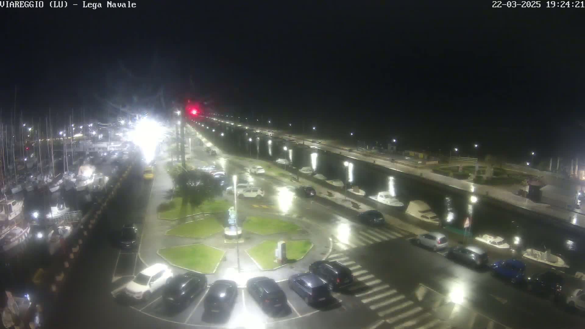 A nighttime, rain-soaked marina with numerous boats docked and cars parked in a lot next to a canal.