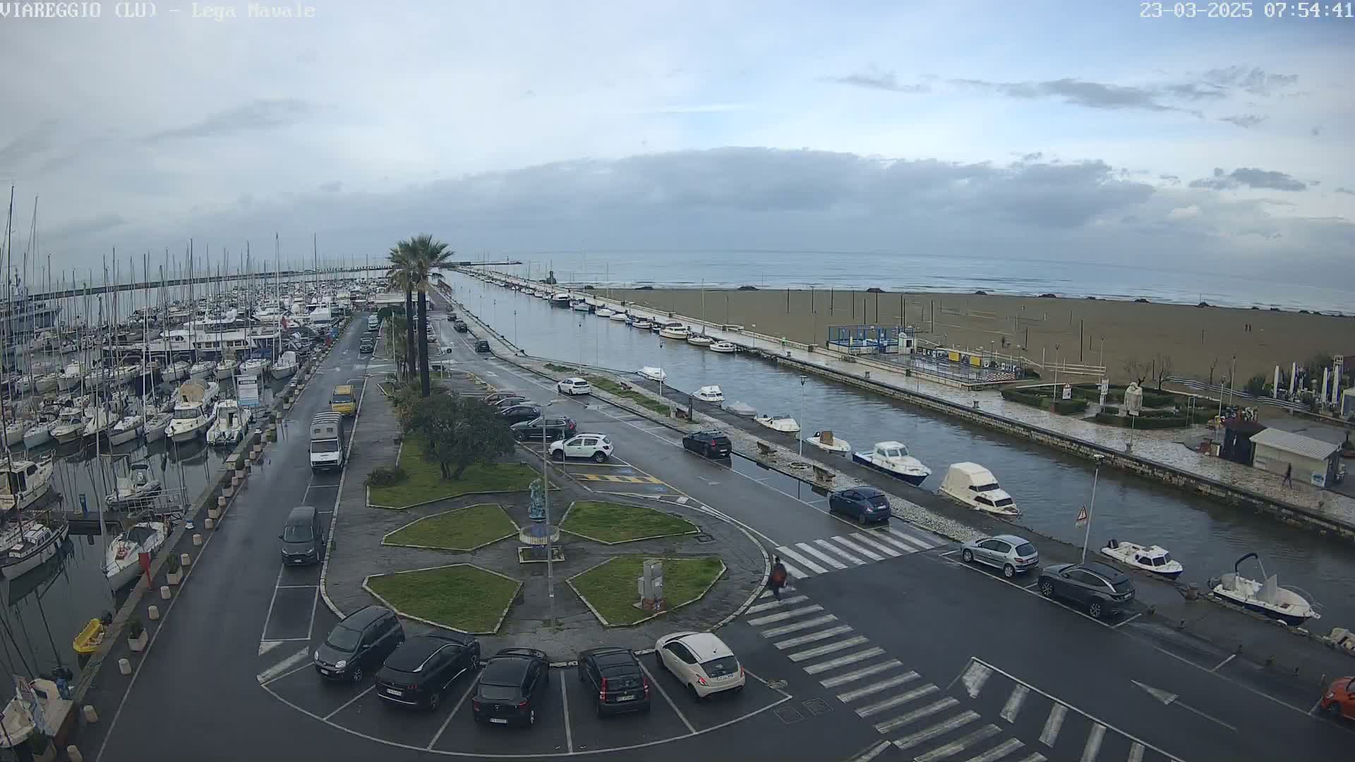 A harbor filled with boats is next to a canal and a beach under an overcast sky.