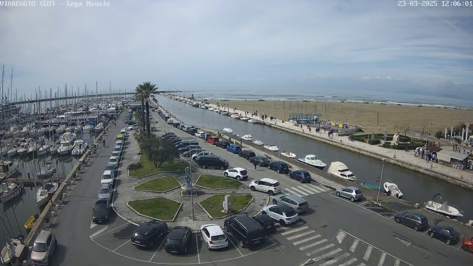 A marina filled with boats and a parking lot next to a canal, all overlooking a sandy beach and ocean under a mostly clear sky.