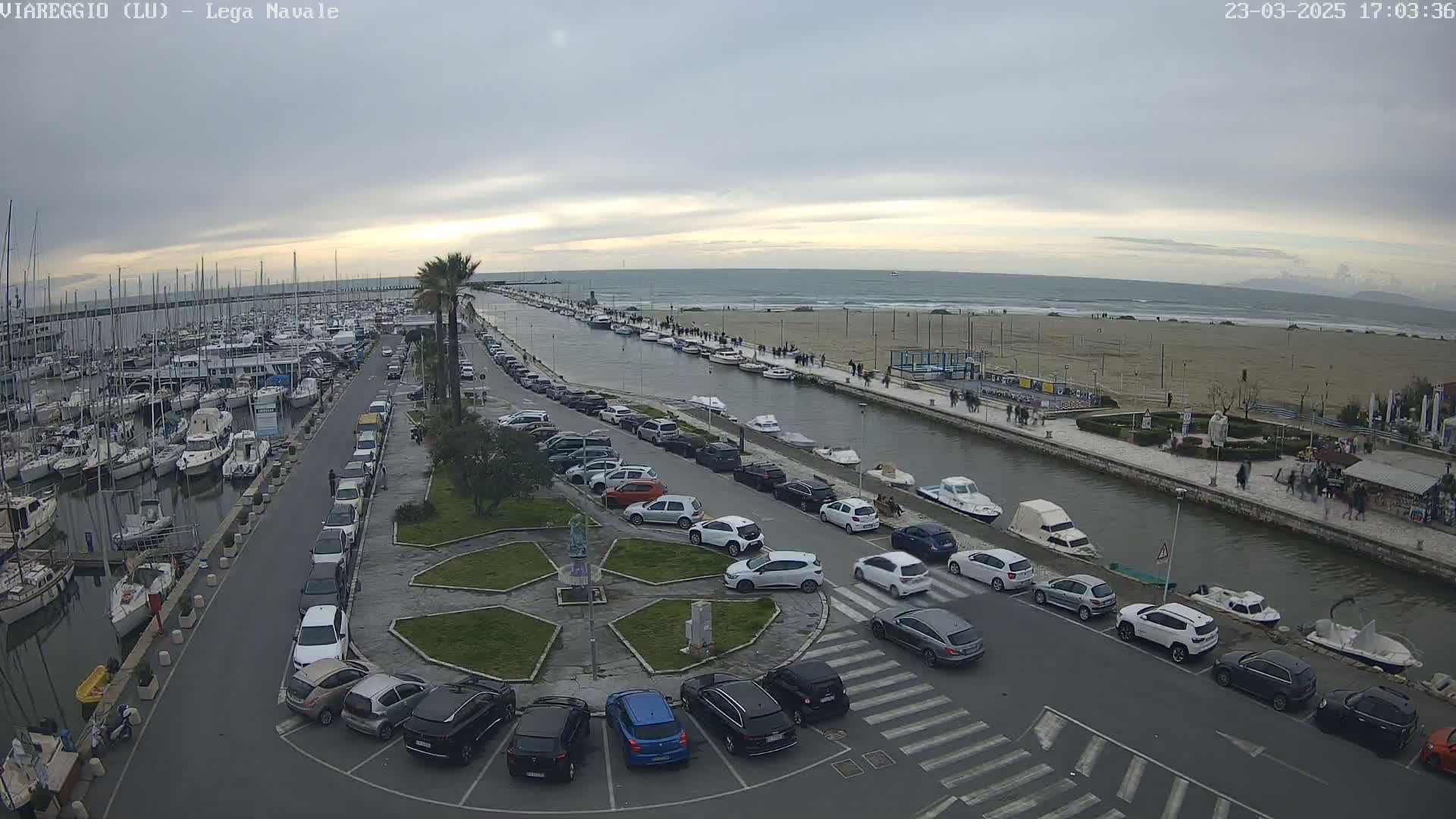 A harbor filled with boats is next to a canal lined with parked cars and pedestrians, all under an overcast sky.