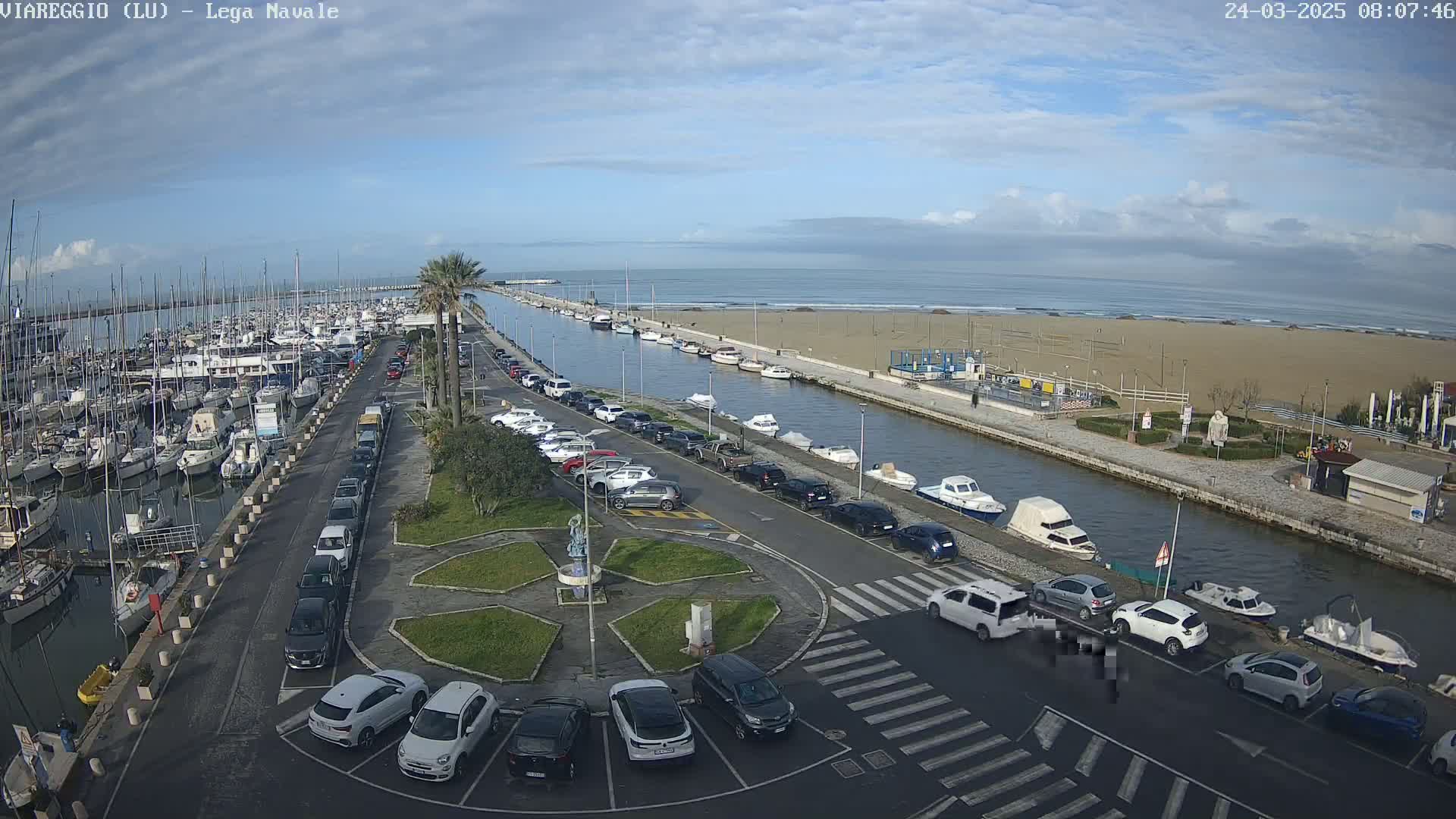 A marina filled with boats is adjacent to a sandy beach and calm ocean under a mostly cloudy sky.