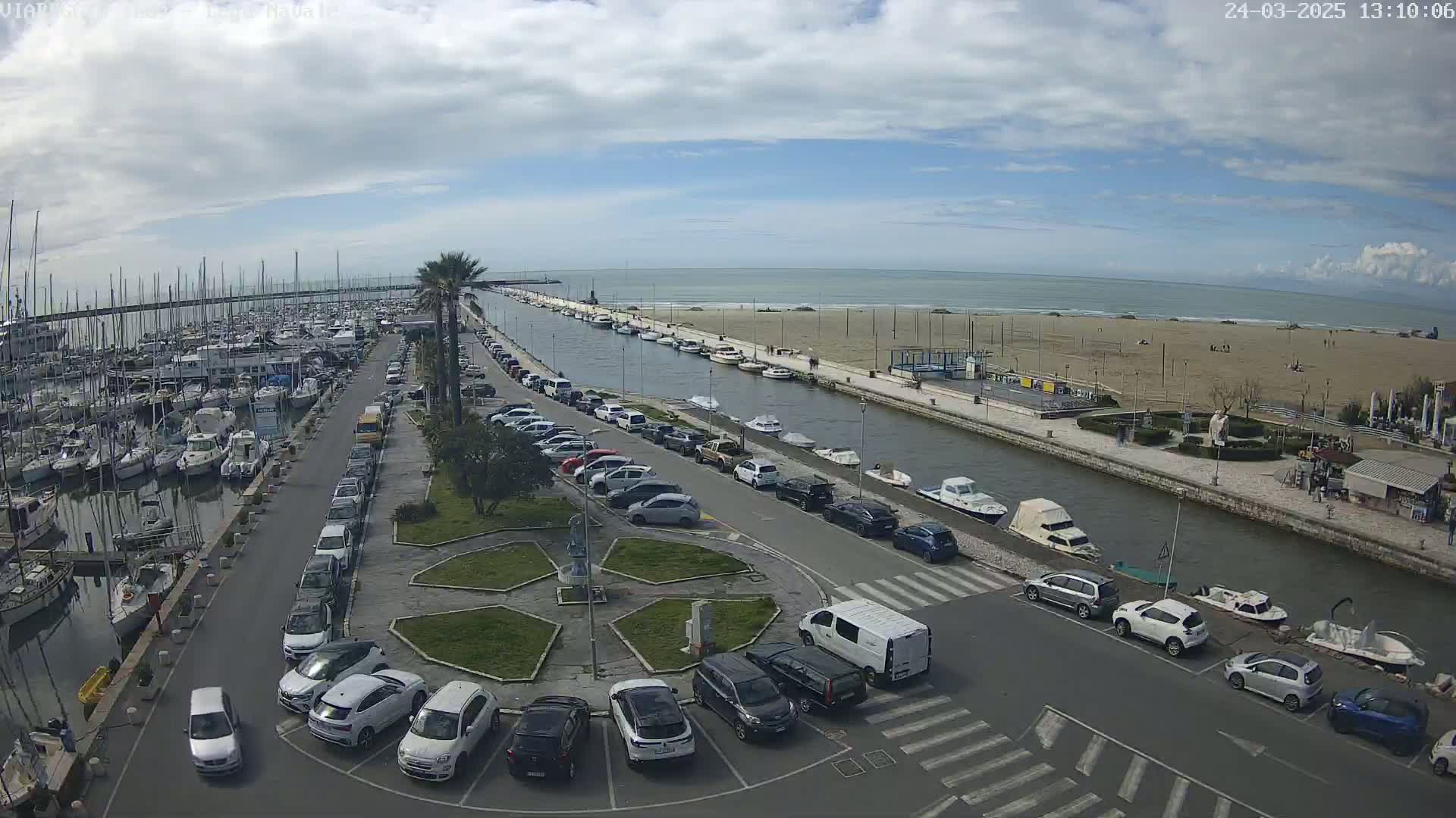 A harbor filled with boats and a parking lot next to a canal leading to a sandy beach under a partly cloudy sky.