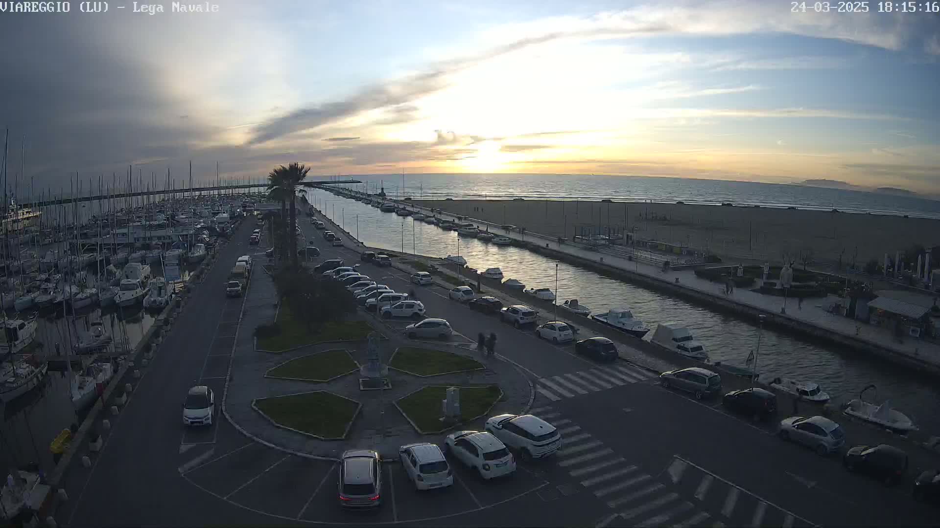 A marina filled with boats is next to a canal leading to a sandy beach at sunset under a partly cloudy sky.