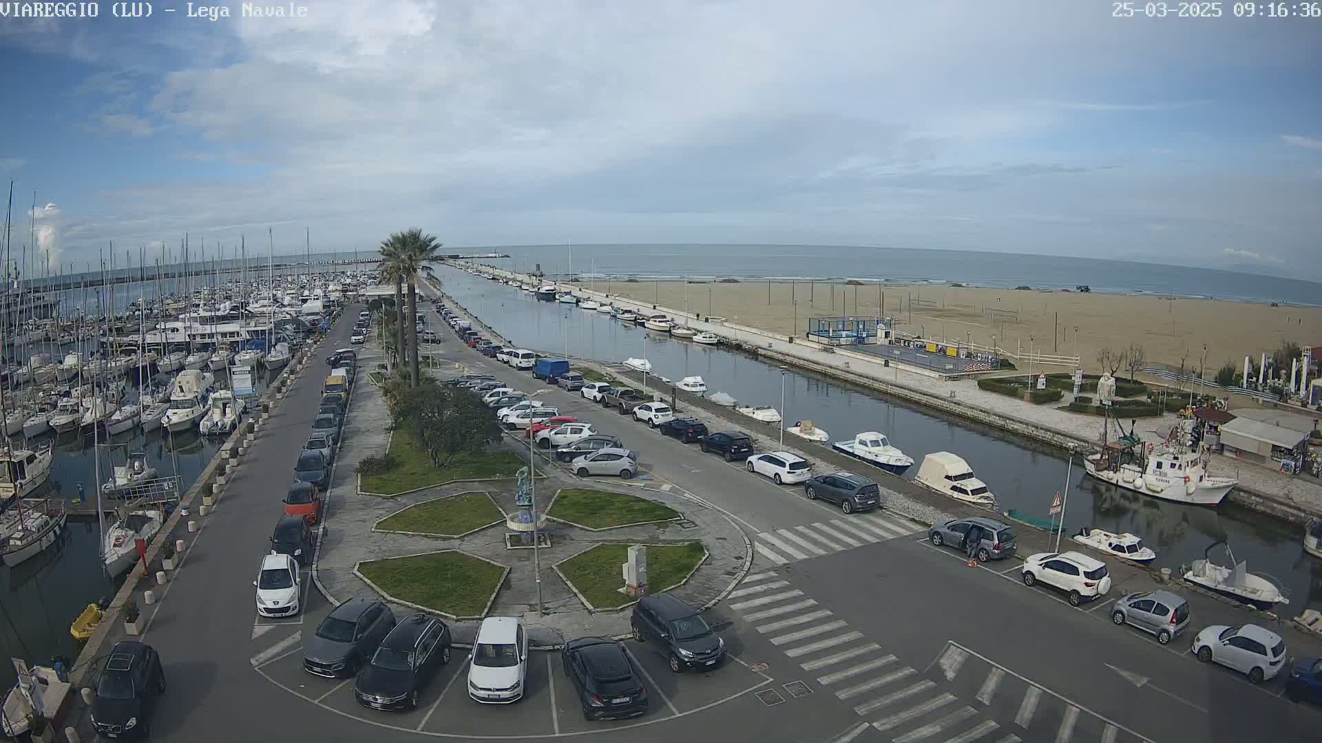A marina filled with boats is adjacent to a canal that leads to a sandy beach and ocean under a partly cloudy sky.