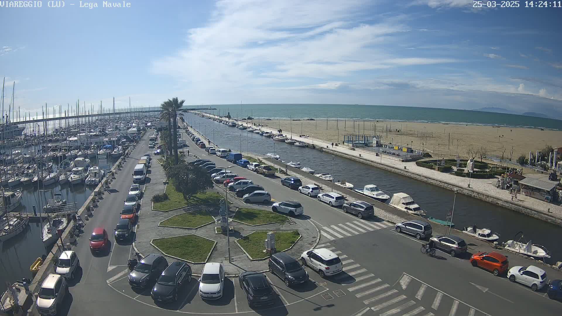 A marina filled with boats is adjacent to a canal that leads to a sandy beach and calm ocean under a partly cloudy sky.