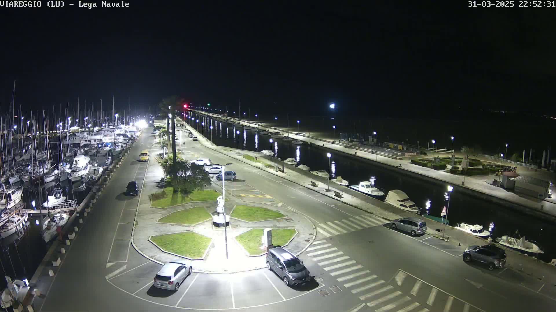 A nighttime aerial view of a marina with numerous boats docked, a paved area with cars parked, and a central roundabout, all illuminated under clear skies.