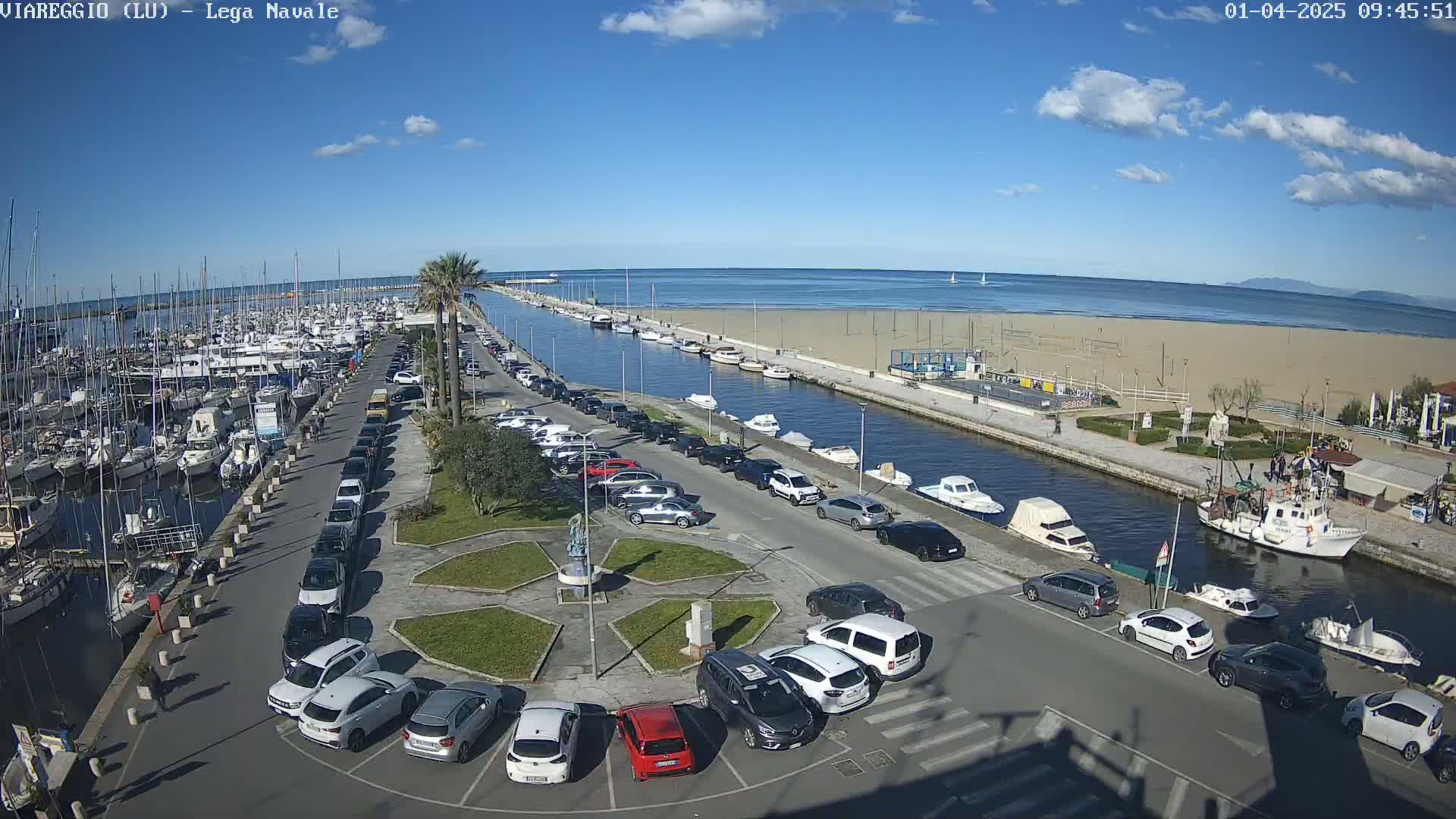 A marina filled with boats is adjacent to a sandy beach and calm ocean under a mostly sunny sky, with a parking lot and canal separating the two.